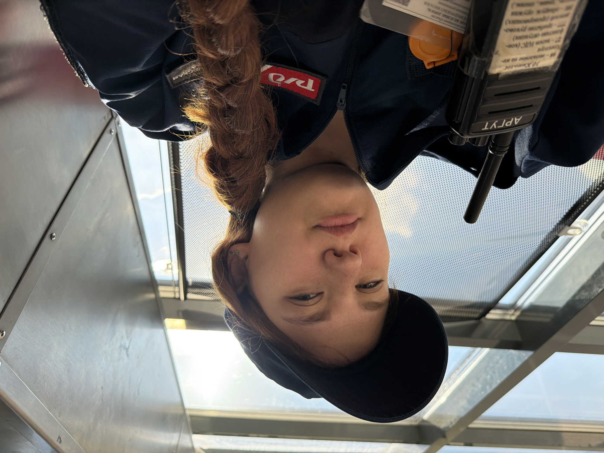 In Rostov-on-Don, Russia, a young woman with red hair braided down her right side is taking a selfie. She is wearing a dark blue baseball cap and a dark blue zippered jacket with a red patch that reads "РЖД" on the left side of her chest. A black walkie-talkie is clipped to the left side of her jacket. The background consists of angled glass panes with a textured screen behind them, suggesting an indoor space with large windows, possibly an observation deck or a transportation hub like a train station. Sunlight streams in from the upper right, illuminating her face and hair. The overall impression is of a person in uniform, possibly on duty, against a modern architectural backdrop.
