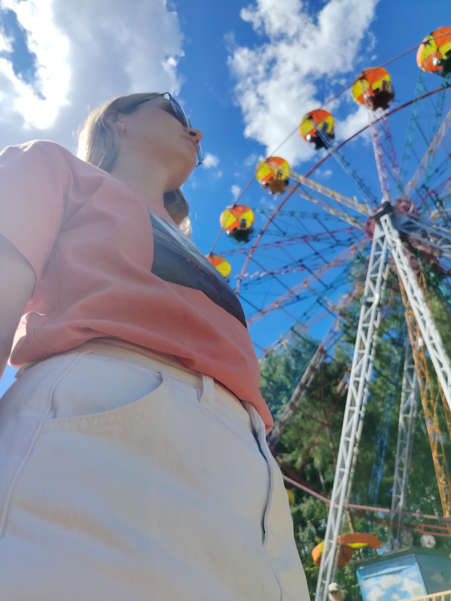 The image shows a low-angle, wide shot of a woman looking up at a Ferris wheel on a sunny day. The woman is in the foreground, wearing a peach-colored t-shirt and light-colored shorts. Her face is partially obscured by sunglasses, and her blonde hair is visible. The Ferris wheel is in the background, with several gondolas visible against a bright blue sky with white clouds. Trees are visible behind the Ferris wheel. The setting appears to be an amusement park. The image was likely taken in the city of Chaikovsky, Russia, as suggested by the location prompt.