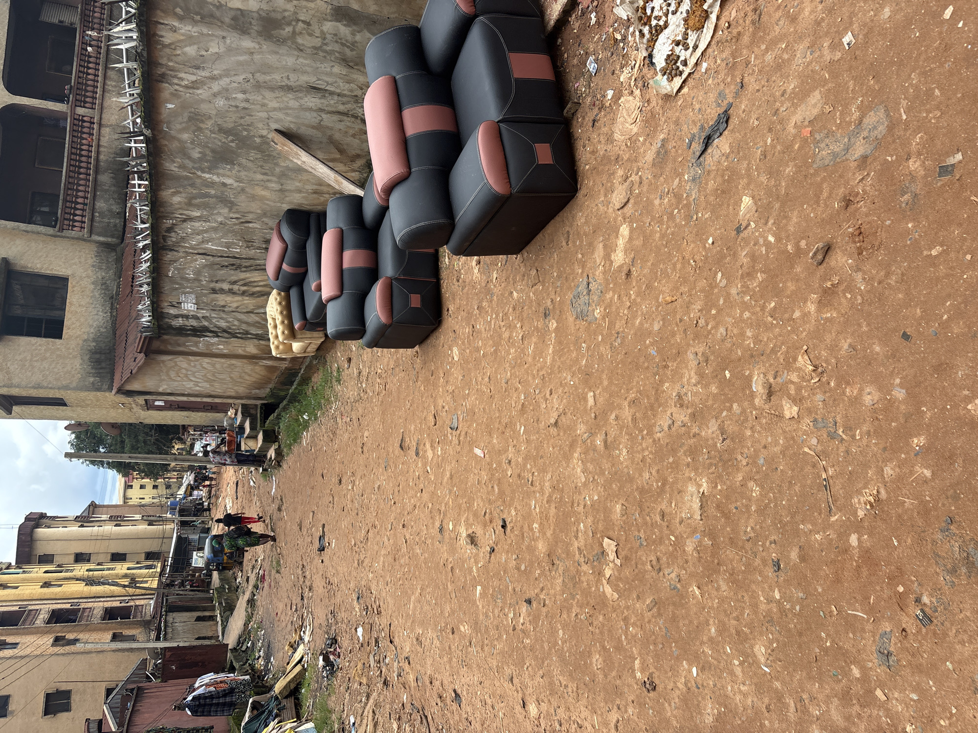 In Onitsha, Nigeria, a narrow, unpaved street is lined with buildings on either side, exhibiting varying states of repair. The street itself is composed of reddish-brown dirt and is littered with debris, including plastic bags, paper, and other refuse. On the right side of the street, several upholstered armchairs, black with pink accents, are arranged along a low wall. A yellow armchair is also visible behind them. The wall is topped with barbed wire fencing. In the distance, several people are walking along the street, and a three-wheeled vehicle is present. The buildings are predominantly multi-story structures with balconies and visible electrical wiring. The sky above is overcast.