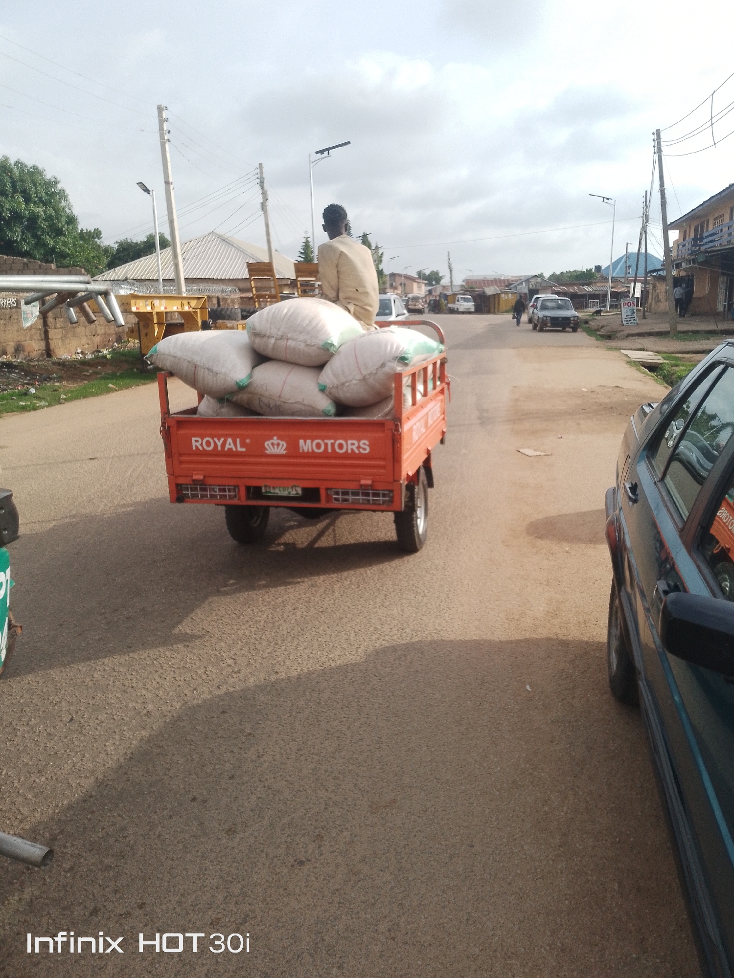 A man in a light-colored shirt and pants sits atop a pile of white sacks in the back of an orange three-wheeled vehicle on a paved road. The vehicle has "ROYAL MOTORS" written on its side. To the right, the side of a dark green car is visible. The background shows buildings, trees, power lines, and streetlights under a cloudy sky. The scene appears to be in an urban or semi-urban area.