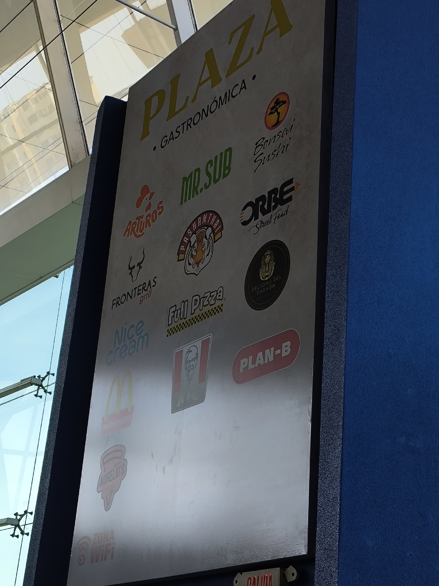 The image shows a sign for "Plaza Gastronómica" with a list of various restaurants and food vendors located in the mall of the same name in the city of Porlamar, Venezuela. The sign lists the following restaurants: Arturo's, Mr. Sub, Bonsai Sushi, Fronteras Grill, Pashamthai, Orbe Street Food, Nice Cream, Full Pizza, Delicias del Faraón, McDonalds, KFC, Aurelio's and Plan-B. Also visible are the words "Zona Wifi" and "Salida". The sign is next to a wall.