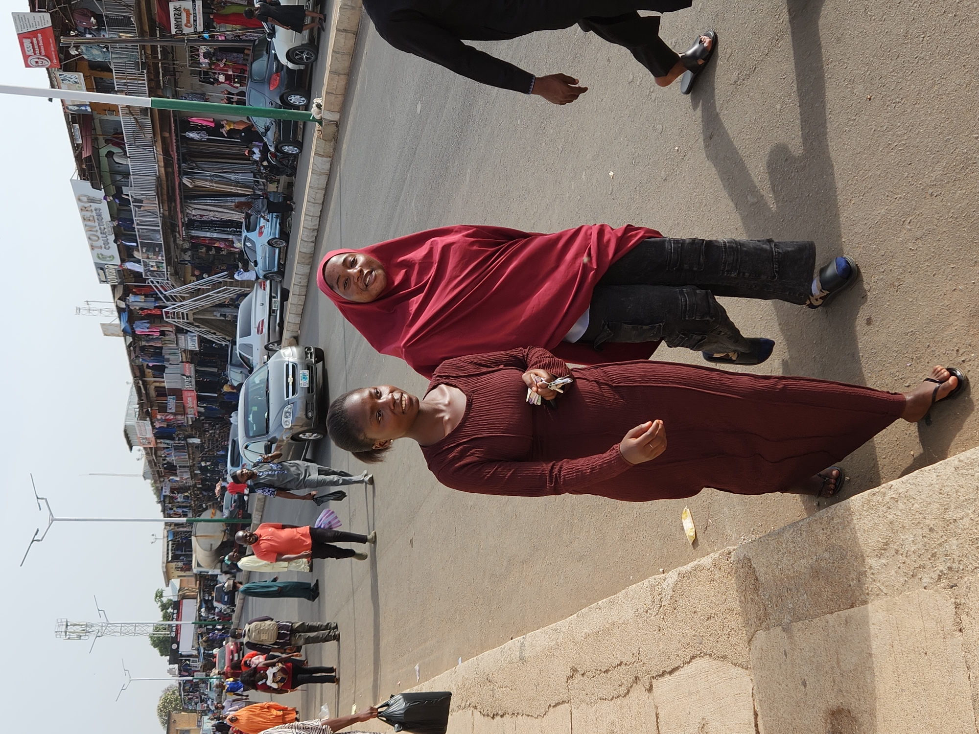 The image depicts a street scene in Jos, Nigeria, showing two women standing on a sidewalk next to a busy street. One woman wears a long brown dress and holds keys in her hand. The other woman is dressed in a red hijab and black jeans. Cars are parked along the street, and there are numerous pedestrians in the background. Shops and buildings line the street, with clothes and merchandise visible on display. Streetlights are also visible.