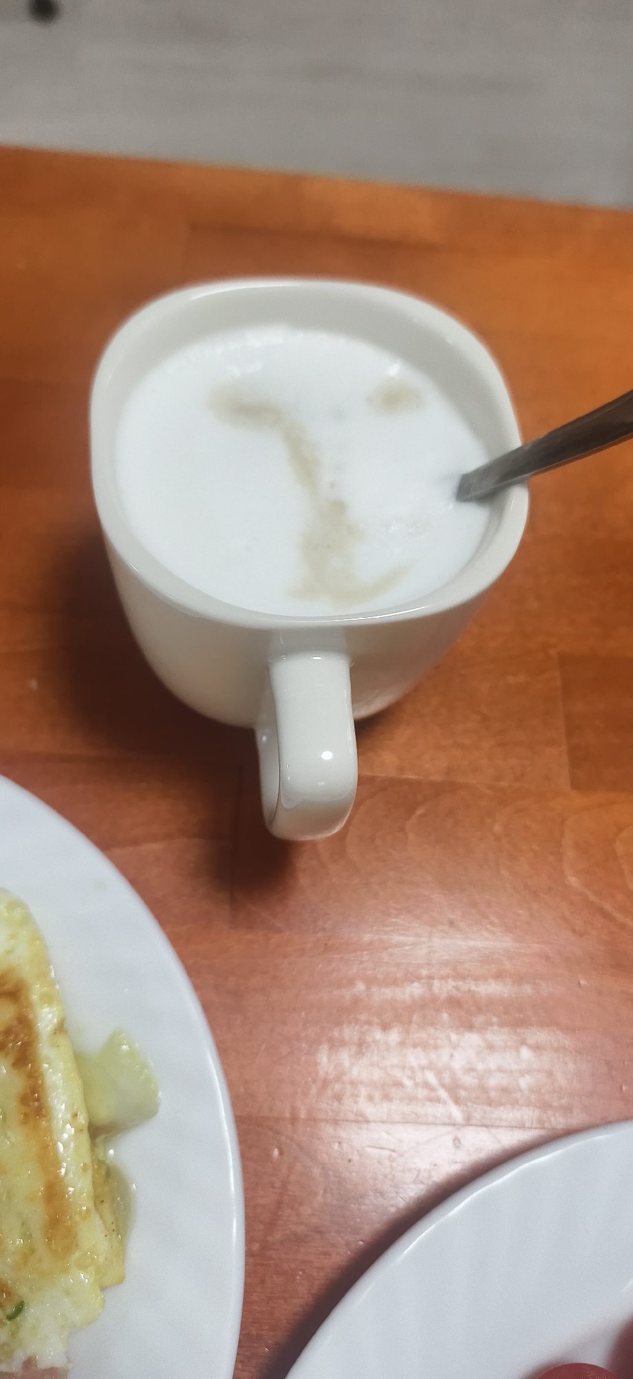 The image presents a top-down, close-up view of a casual breakfast or brunch setting on a wooden table in Fokino, Russia. The main subject is a white ceramic mug, filled with a white, frothy beverage, likely coffee with milk foam, such as a latte or cappuccino. A silver spoon rests within the mug. The milk foam exhibits an abstract, light brown pattern that loosely resembles a stylized letter 'T' or an elongated, indistinct facial shape.
To the left of the mug, a portion of a white plate is visible, holding a serving of what appears to be an omelet or scrambled eggs, with some yellow-green bits mixed in. Another white plate is partially visible in the bottom right corner, containing blurred reddish items that could be sliced tomatoes or small sausages. The table surface is made of warm, light brown wood with visible grain. The lighting is soft and consistent with an indoor environment. No people are visible, and no text is discernible within the frame. The scene conveys a quiet, domestic moment, possibly during the morning, focused on the food and drink.