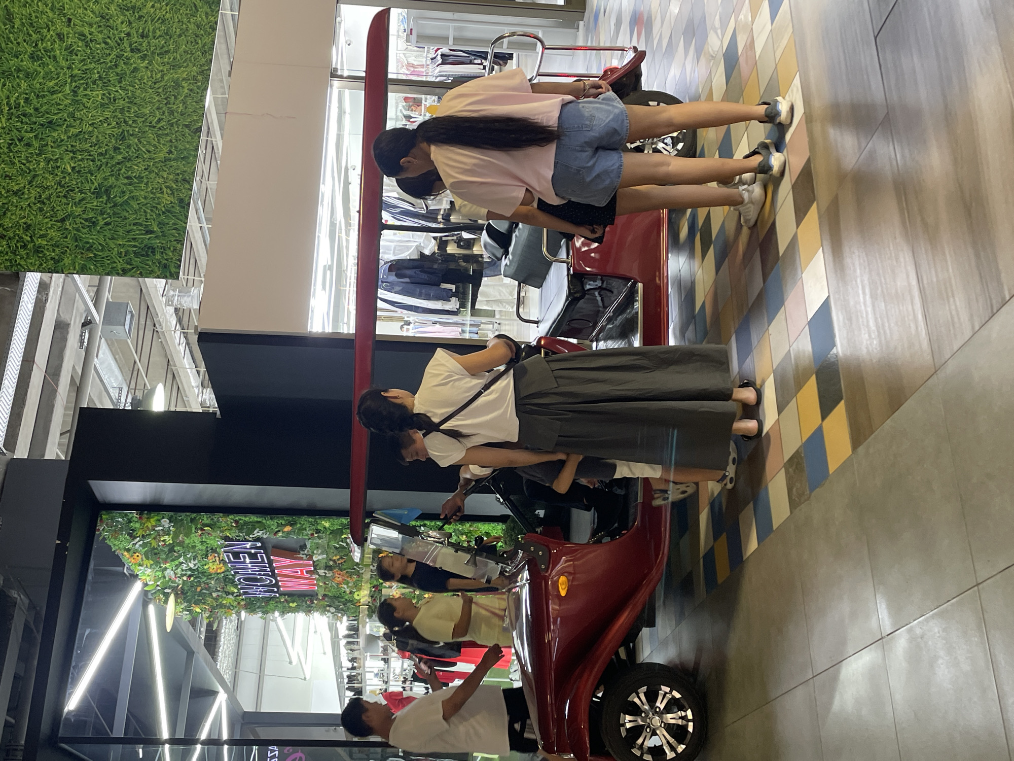 A group of people are gathered around a small, open-air, red vehicle with a black seat and a red canopy, likely in a mall. The vehicle appears to be a recreational or novelty item, possibly a tuk-tuk style buggy. A young woman in a white t-shirt and a long, dark grey skirt stands facing away from the camera, her back to the vehicle. To her right, two young women are also facing away, one in a pink t-shirt and denim shorts, and the other in a white t-shirt and a black skirt. Another person, possibly a child, is partially visible in the driver's seat of the vehicle. To the left of the vehicle, a man in a white t-shirt and dark shorts is looking at his phone, and a woman in a yellow t-shirt and light-colored pants stands beside him. In the background, there is a storefront with mannequins dressed in clothing and a large decorative wall covered in artificial plants and flowers, with the words "WOMEN WAY" illuminated in neon pink letters. The floor is tiled with a checkered pattern of various colors. The ceiling appears to be a concrete structure with exposed beams and lighting fixtures. The overall impression is a retail or entertainment environment. The specific location of Almaty, Kazakhstan is not discernible from the image itself.