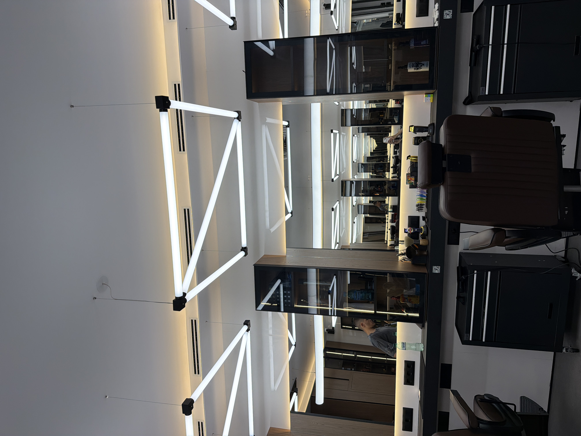 This image depicts the interior of a modern barbershop, likely located in Sofia, Bulgaria. The space is brightly lit by a network of geometric, white fluorescent tube lights suspended from the ceiling, creating a contemporary and clean aesthetic. Several barber chairs with brown leather upholstery are visible, positioned in front of mirrored stations. One station features a man, possibly a barber, who appears to be working or inspecting something. Behind the stations, shelves stocked with various barbering products and equipment are visible through glass cabinets. The overall impression is of a well-equipped and stylish establishment designed for professional haircuts and grooming.