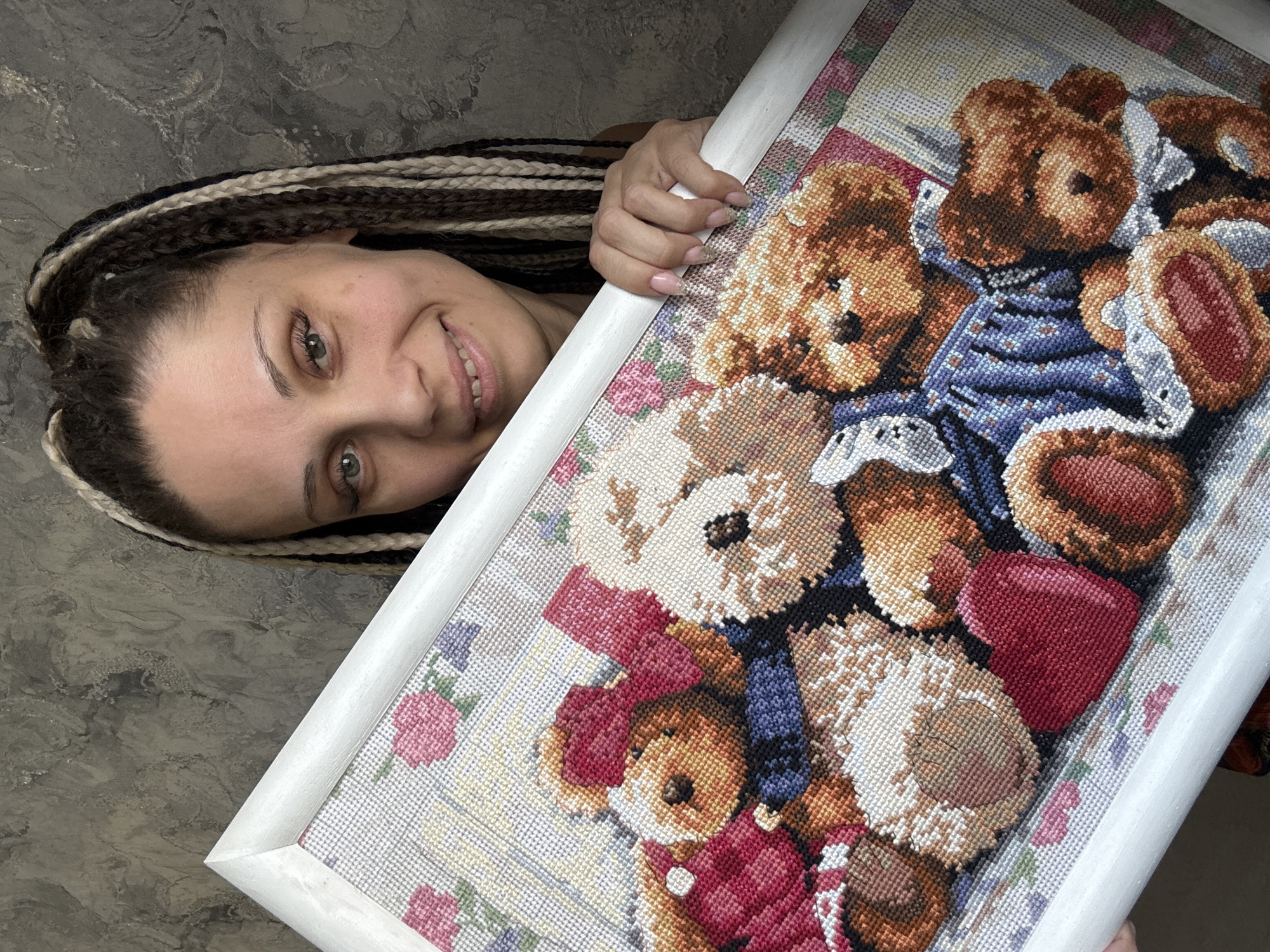 A woman with a gentle smile and light-colored eyes is captured holding a framed cross-stitch artwork. Her head is tilted, and she looks towards the viewer with a pleasant expression, seemingly presenting the piece. She has distinctive braided hair, featuring a mix of dark and light strands.
The main subject of the image is the cross-stitch artwork, which depicts a cozy group of teddy bears. Several bears are visible, rendered in various shades of brown and beige, showcasing the intricate detail of the needlework. One prominent teddy bear in the upper part of the frame wears a blue patterned outfit, while another near the bottom left is adorned in red with a matching bow. The background of the cross-stitch features subtle floral and geometric patterns. The artwork is enclosed in a simple, white wooden frame.
The woman's hand, with naturally manicured nails, is visible gripping the frame. Behind her and the artwork, there is a textured, mottled gray surface, likely an indoor wall. The overall mood conveyed by the woman's smile and the artwork's subject matter is one of warmth, pride, and domesticity. The image appears to be well-lit. There is no visible text within the photograph. Based on the provided context, the location is Rusanovo, Russia.