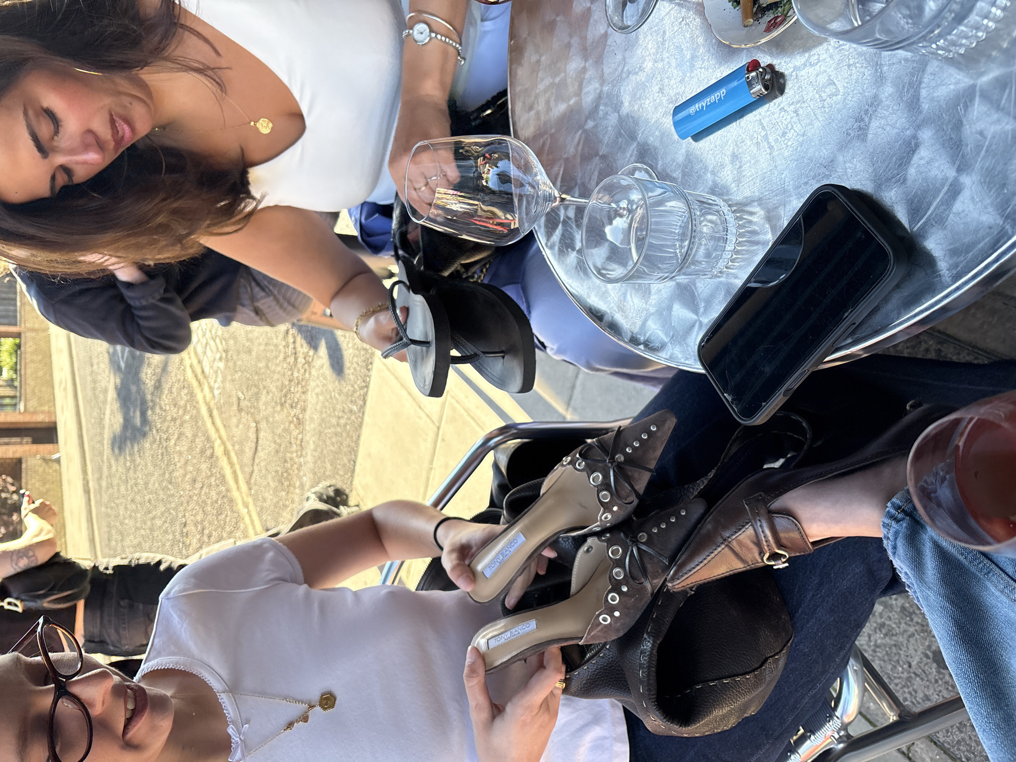 This is a medium shot photograph taken outdoors on a sunny day, likely in London, United Kingdom, as indicated by the architecture visible in the background. The focus is on a group of people seated at a table, with the foreground occupied by two women and various objects on the table.
One woman, on the left, is wearing glasses and a white t-shirt with a delicate necklace. She is smiling and appears to be in conversation. The other woman, on the right, is wearing a white top and a gold necklace with a coin pendant. She is looking down at the table and holding a pair of black flip-flops in her right hand. Her left hand is resting on the table. She also has a watch on her left wrist.
On the table, there are several objects: a wine glass filled with red liquid, an empty glass, a smartphone in a black case, a blue lighter with white lettering, and a small ashtray with cigarette butts. Two pairs of shoes are also visible, one pair of brown stiletto heels with silver embellishments held by the woman on the right, and another pair of brown pointed-toe flats on the lap of the woman on the left. The table itself is a circular metal table with a brushed metallic surface.
In the background, more people are visible, suggesting a public setting like a cafe or restaurant patio. The lighting indicates it is late afternoon or early evening, with strong shadows cast by the sunlight. The overall scene suggests a casual social gathering.
