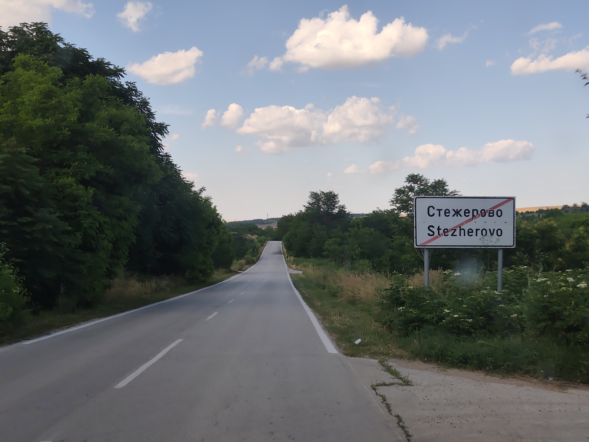 The image depicts a road sign for "Стежерово Stezherovo" in Bulgaria. The sign is white with red lettering and a red diagonal line across it, indicating the end of the town. The road is asphalt and curves to the left, leading into a rural landscape with trees and vegetation on either side. The sky is blue with scattered white clouds. The setting appears to be during the daytime, likely late afternoon due to the warm light. There are no people or vehicles visible in the image. The overall scene is one of a rural road entering or exiting a small Bulgarian town.