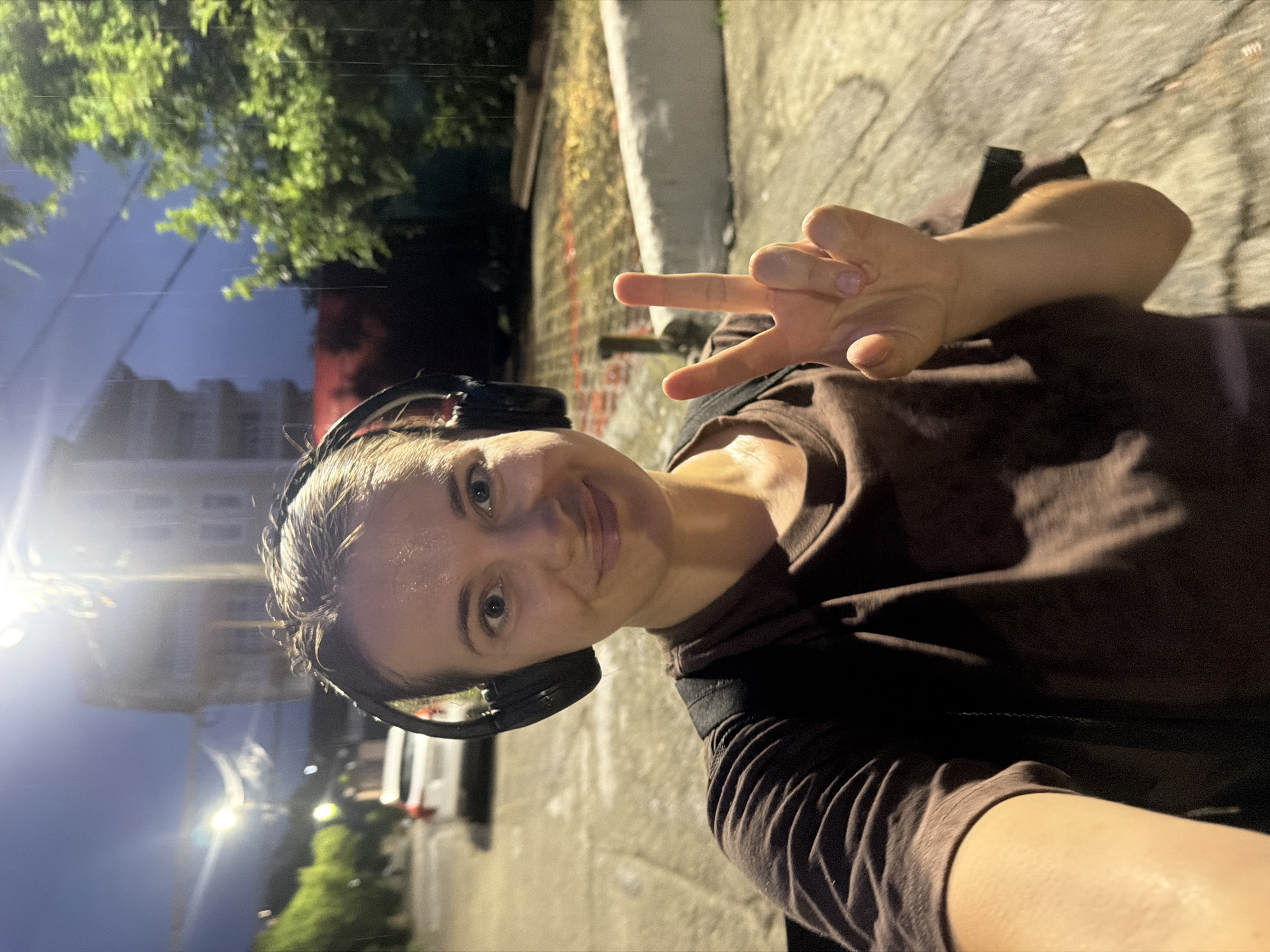 In a selfie taken outdoors at night, a woman with headphones on holds up two fingers in a peace sign. The woman is wearing a brown t-shirt and appears to be sweating. The background shows a street with a car parked and a building in the distance. Raindrops are visible in the image, indicating it is raining. The lighting is dim, with a street lamp visible in the upper portion of the image. The woman's expression is a smile. The location is not identified as Sochi, Russia.