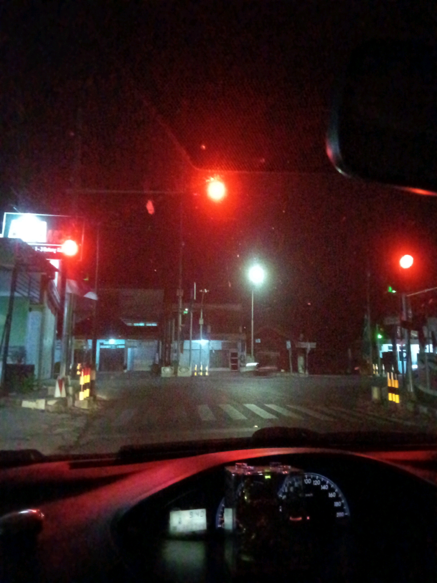 The image shows a view from inside a car at night, looking out the windshield. The car appears to be stopped at a traffic light or toll booth in Ngasem, Indonesia. Red traffic lights are visible in the distance, illuminating the dark road. Buildings and streetlights are also present, creating a dimly lit urban environment. The car's dashboard is visible in the foreground, with a speedometer showing a speed of 0 km/h, indicating the car is stationary. The scene is dark and somewhat grainy, typical of nighttime photography.