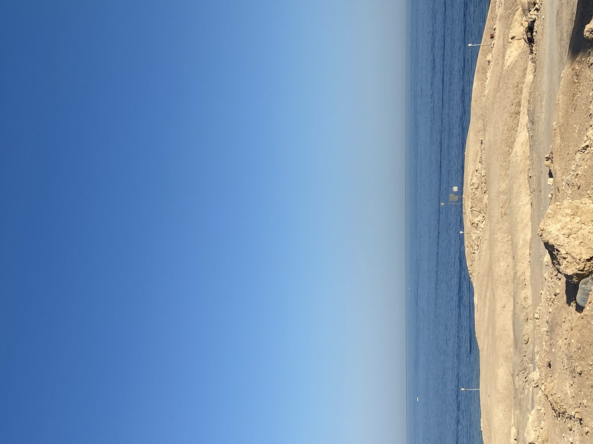 The image shows a vast expanse of blue sky meeting the calm blue sea. The horizon line is clearly defined, with the sky gradually lightening towards it, suggesting a bright, clear day. The water is a deep, consistent blue with very subtle ripples.
On the right side of the image, a rocky, sandy embankment rises from the water's edge. The embankment appears dry and arid, with a rough, textured surface. A paved path or road runs along the top of the embankment. Several lampposts are visible along this path, some with a red cylindrical object at their base. The presence of these lampposts suggests this may be a coastal road or promenade. In the far right of the image, a plastic water bottle is visible on the rocky ground.
The overall scene depicts a serene coastal environment. The clear sky and calm sea suggest pleasant weather. The time of day appears to be daytime, likely late morning or early afternoon, given the bright lighting. The location cues of arid land meeting the sea, along with the presence of lampposts, are consistent with a resort or coastal area. There is no discernible activity or event taking place; the image captures a still, natural landscape. No text is visible in the image.