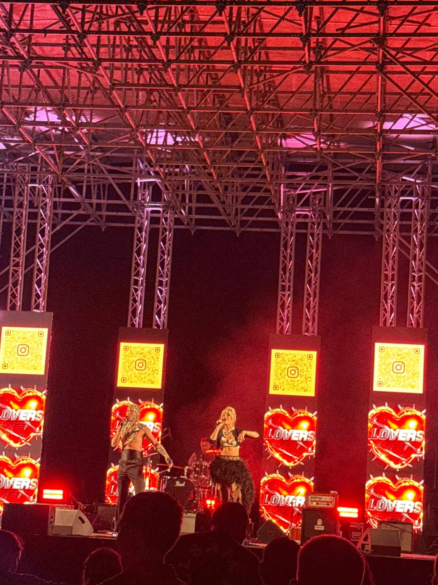 The image depicts a concert stage in Limassol, Cyprus, likely at the Limassol Marina. The stage is adorned with a complex metal truss structure illuminated by red and pink lights. Large screens display glowing red hearts with the word "LOVERS" and Instagram QR codes. Two female performers are on stage: one in the center, wearing a sparkly hooded top and black pants, appears to be singing; another on the left, in a black outfit with a feathered skirt, is also singing. A drum kit and amplifiers are visible on stage. The audience, mostly out of focus, is seated in front of the stage.