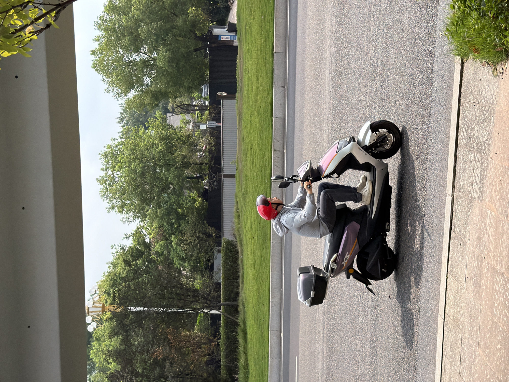 A person wearing a gray hooded jacket and dark pants, along with a red helmet, rides a silver and purple electric scooter with a black storage box on the back. The scooter is on a paved road with a white dividing line. In the background, there is a well-maintained green grassy area with trees and a dark fence. The scene appears to be in an urban or suburban setting, possibly in Jiangjin, China, given the context. The sky is overcast. The foreground shows a pedestrian walkway with light-colored square paving stones and some greenery.