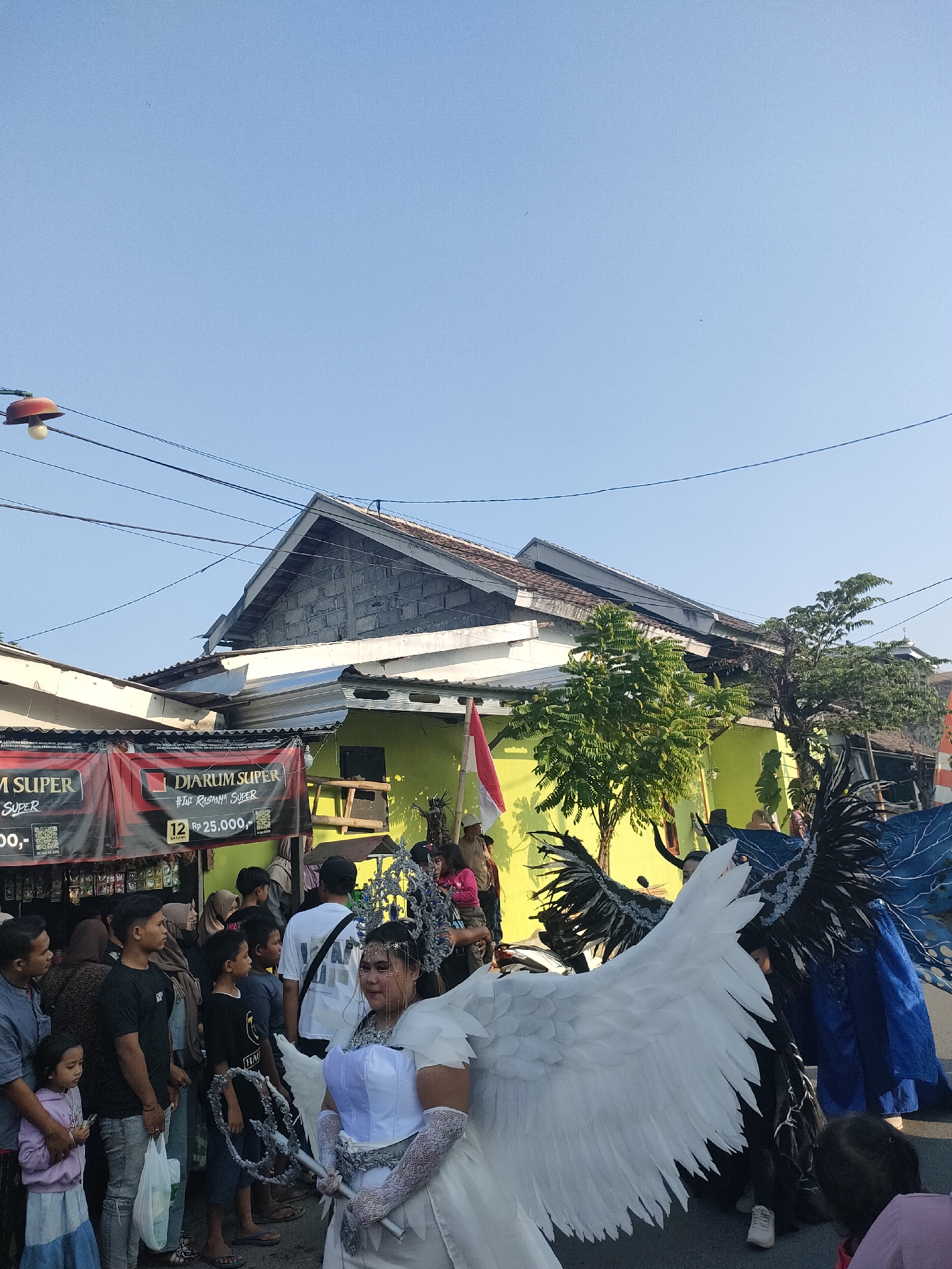 In the city of Tongaswetan, Indonesia, a street procession is underway. The scene is dominated by a person in a white, angelic costume with large feather wings, holding a silver staff and wearing an ornate headpiece. To their right, another individual is adorned with dark, intricate wings. A crowd of onlookers, a mix of adults and children, lines the street, some holding bags and phones. Behind the procession, a lime green building is visible, with a tiled roof and a red and white Indonesian flag draped from its side. Banners advertising "Djarum Super" are displayed, suggesting a commercial sponsorship for the event. Power lines crisscross the bright blue sky above. The atmosphere appears festive, with people gathered to witness the parade.