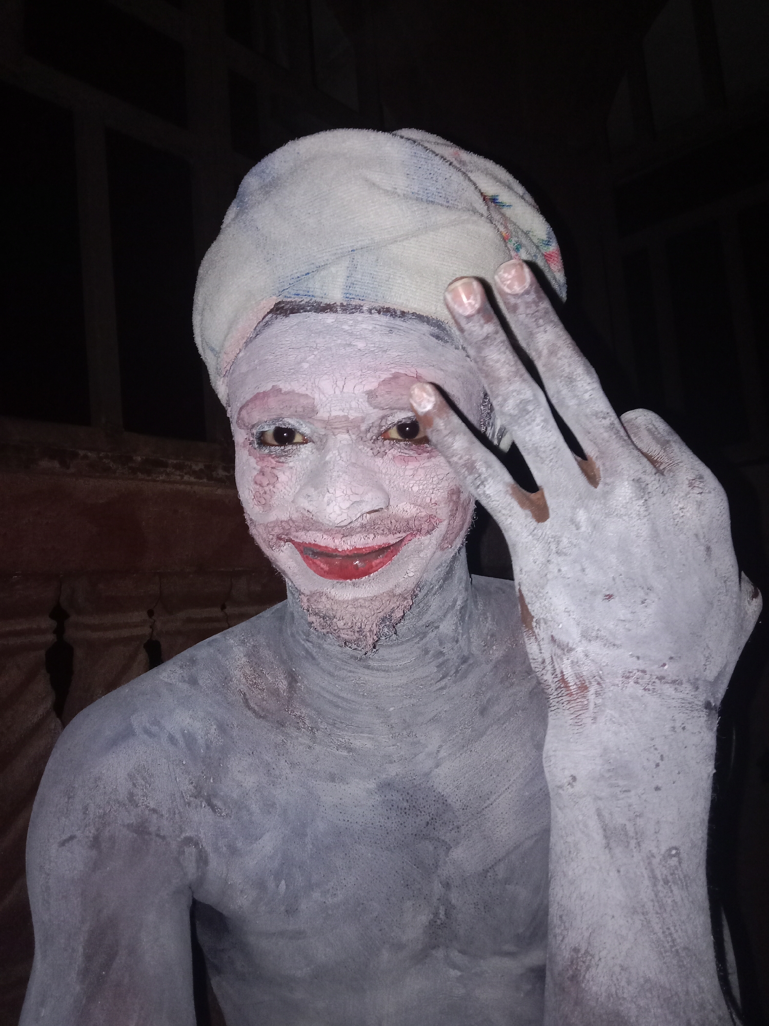 A person covered in white clay and red paint smiles and holds up a hand in front of a dark background. The person is wearing a white towel wrapped around their head. The location is inferred to be Obosi, Nigeria.