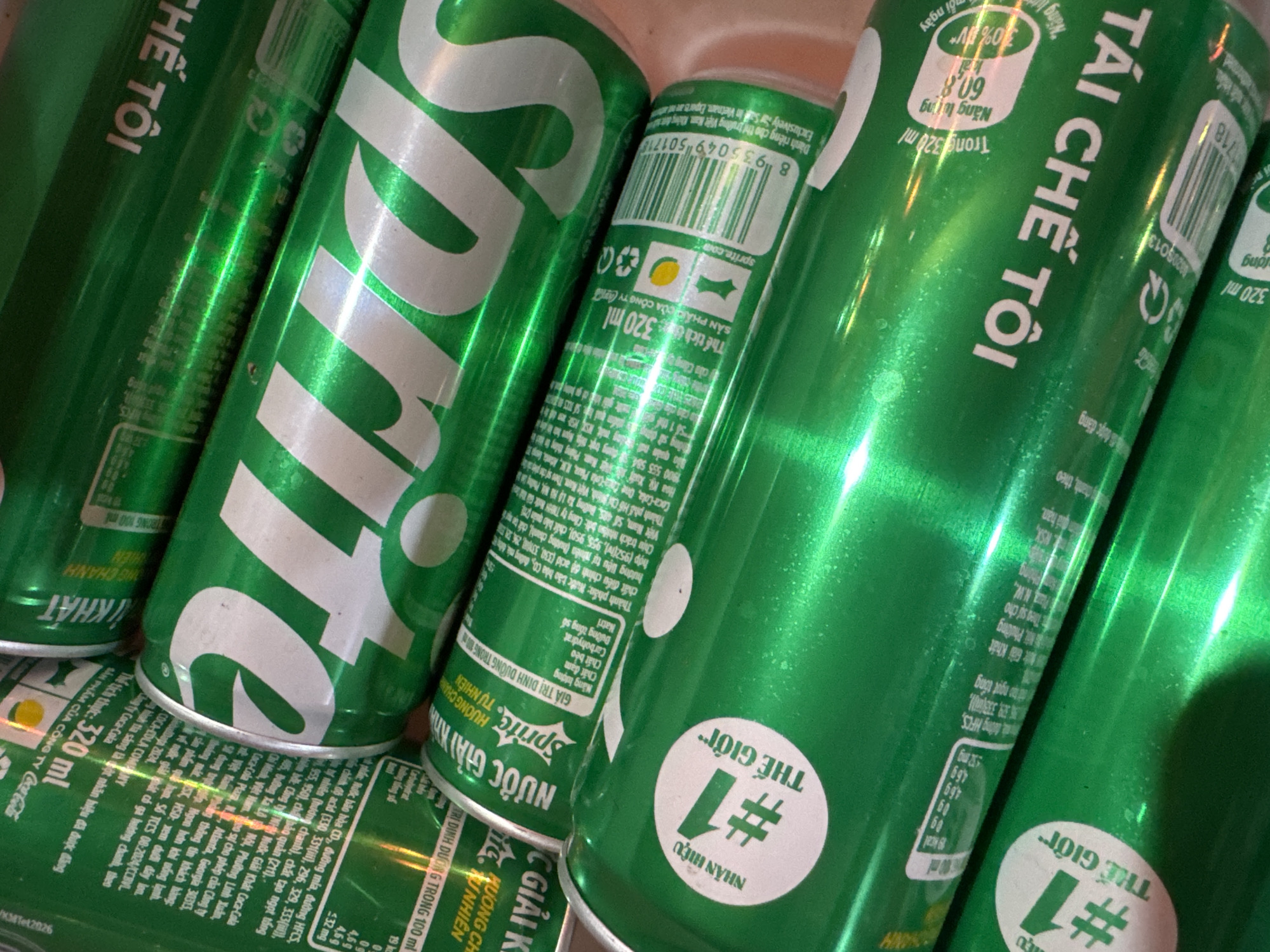 The image shows a close-up of several cans of Sprite soda. The cans are green with white lettering. Some cans display the barcode and nutritional information in Vietnamese. A round sticker on some cans says "#1 Thế giới" (World's #1 Brand). The cans are positioned close together with some cans overlapping others. No people, actions, or locations (including the specified Mèo Vạc Commune, Vietnam) are visible in this specific image. The environment appears to be indoors, with the cans possibly stored in a container or placed on a surface.