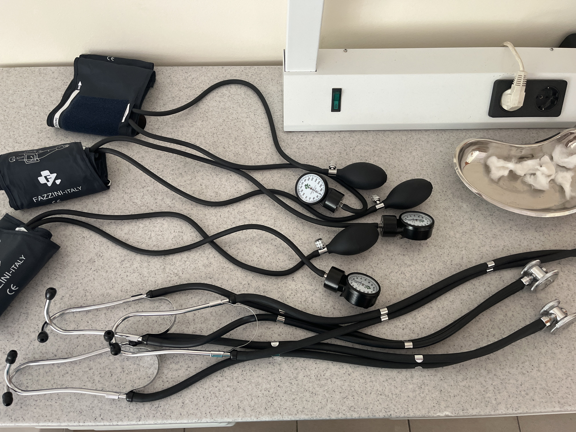 The image shows a medical examination table in an unknown city. Three stethoscopes and three sphygmomanometers are laid out on the table. A kidney dish containing cotton balls is also visible. An electrical outlet and switch are mounted on the wall behind the table.