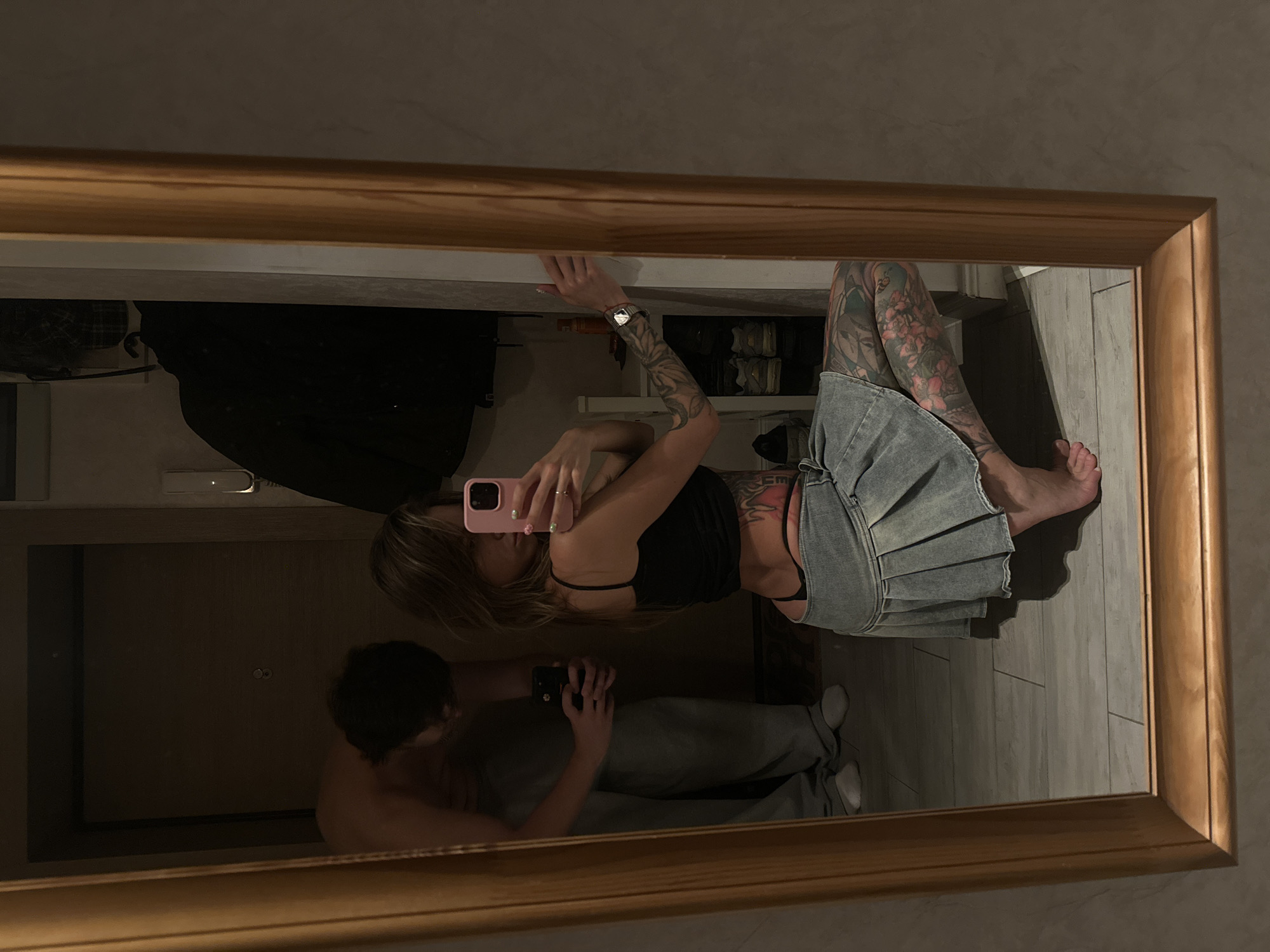 In a dimly lit apartment hallway in Moscow, Russia, a woman crouches in front of a full-length mirror, taking a selfie with her pink phone. She wears a black tank top, a denim mini-skirt, and a black thong, with extensive tattoos visible on her legs and torso. Behind her, a man, shirtless and wearing grey sweatpants, stands and looks at his own phone. The hallway floor is tiled, and a wooden-framed mirror reflects the scene. A closet with hanging clothes and shelves of shoes is visible in the background.