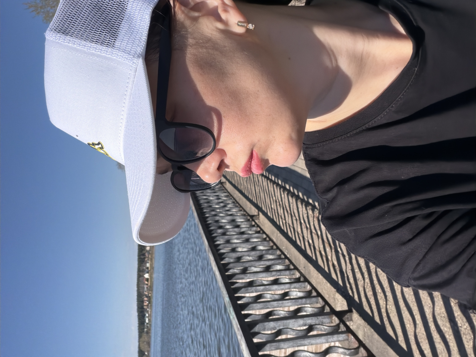 A person wearing a white baseball cap and black sunglasses stands next to a railing. The railing overlooks a body of water. On the opposite shore are buildings and trees. The person is wearing a black long-sleeved shirt and has an earring in their left ear. Based on the location, this is likely in Votkinsk, Russia, overlooking the Votkinsk Reservoir.