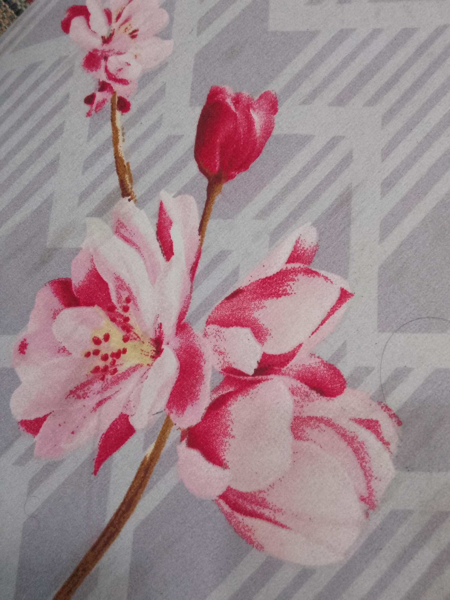 The image displays a close-up, overhead view of a floral pattern on fabric. The pattern features several pink and white flowers with red accents, along with thin brown stems. The background of the fabric has a subtle grey geometric design composed of diagonal lines. There are no people or environmental elements indicative of a specific location in Karangan, Indonesia, or any other city. The scene is solely focused on the textile design.