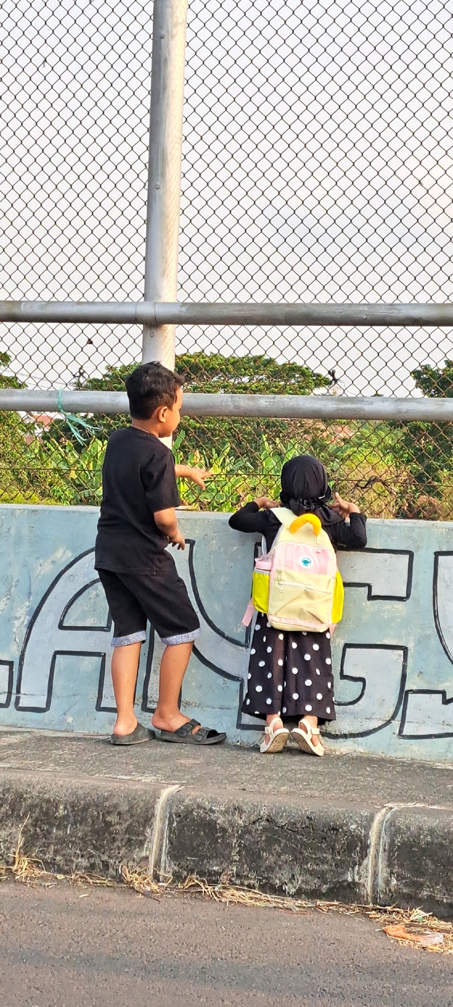 In the city of Kebon Waris, Indonesia, two children stand by a chain-link fence, looking at something beyond it. The boy on the left, wearing a black t-shirt and shorts, points forward with his left hand. The girl on the right, in a black hijab, a polka-dot dress, and a light yellow backpack, leans against a concrete wall with graffiti. She has her hands near her face, possibly shielding her eyes or pointing. They are positioned on a sidewalk or raised path. In the background, lush green foliage is visible, with trees and plants. The sky is overcast, suggesting it is either early morning or late afternoon.