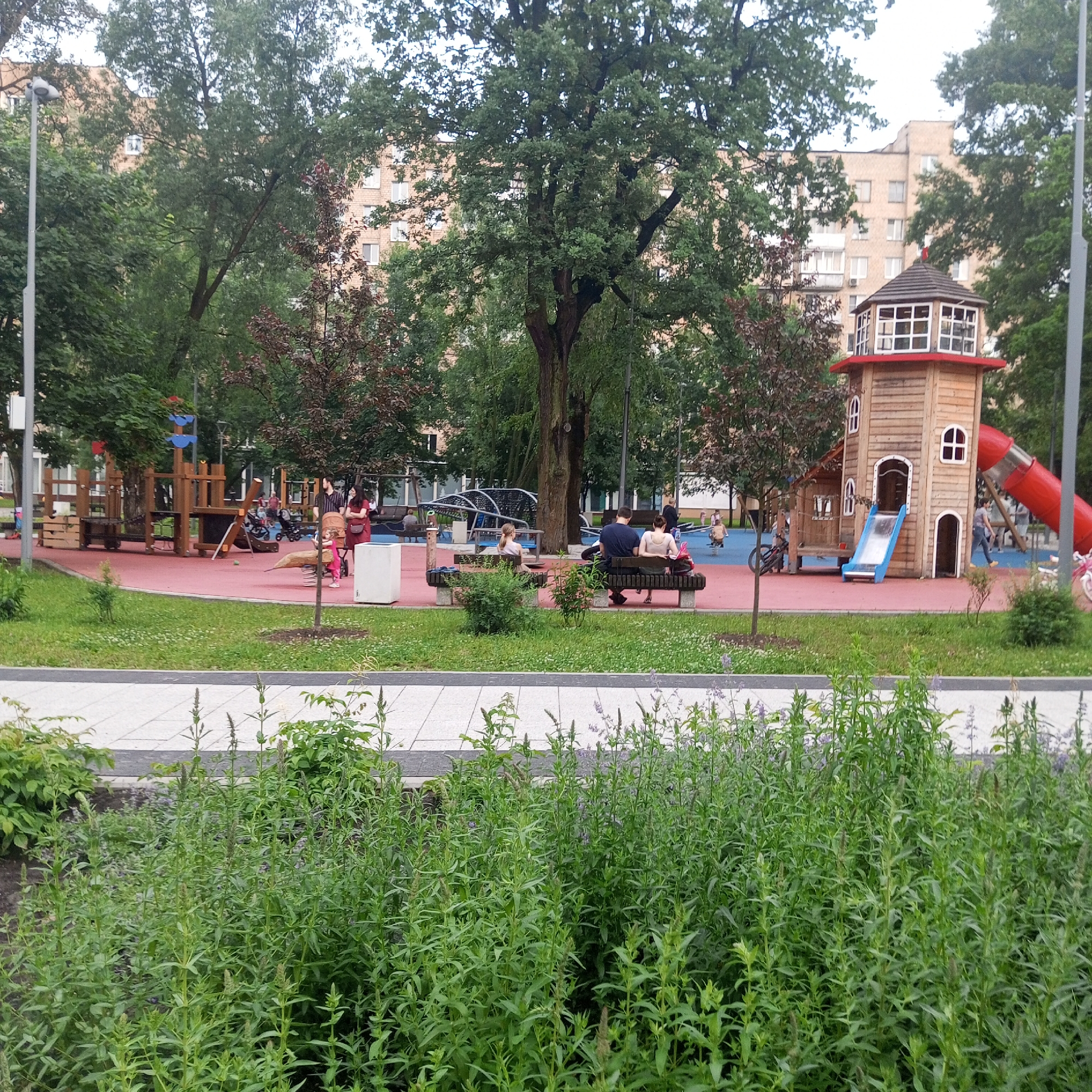 This image depicts a playground area in Moscow, Russia, with a large wooden playhouse resembling a lighthouse, complete with a red slide. In the background, several residential buildings are visible, with trees and greenery surrounding the playground. A group of people, including adults and children, are present in the scene, some sitting on benches and others engaged in playground activities. The foreground is filled with lush green vegetation, suggesting a park or public space within the city.