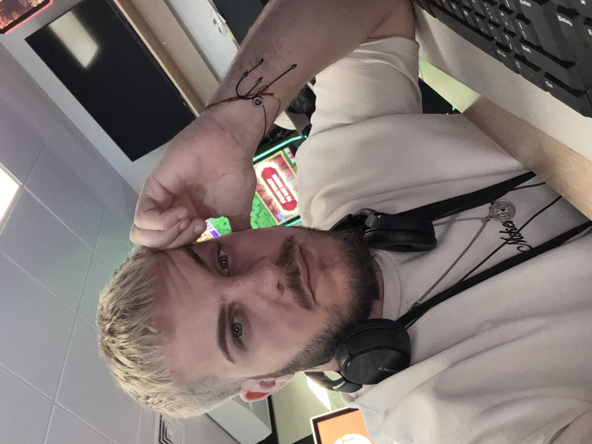The image captures a close-up, rotated portrait of a young man, likely taken as a selfie or by someone nearby. The man has light blonde or ash-blonde hair, a mustache, and a beard. He is wearing a white long-sleeved shirt or hoodie, with black over-ear Sony headphones resting around his neck. A silver chain necklace with a circular pendant, possibly featuring a compass rose design, is visible around his neck. His expression is neutral to slightly serious.
On his right forearm, there are three small, dark shapes resembling stylized fish hooks or anchors, possibly a tattoo or drawn design. A red string bracelet with a small, circular metallic bead is also on his wrist.
The background is somewhat blurry but suggests an indoor setting, potentially a gaming establishment or internet café in Sofia, Bulgaria. Behind him, parts of what appear to be brightly lit screens from gaming or slot machines are visible. On the right side of the frame, a black computer keyboard with visible keys like "Enter," "Delete," and "Ctrl" is on a desk or surface. Another screen in the background shows partially visible text, which includes "PRESS START" on a red background. The ceiling above him features white square tiles and what looks like a ventilation grate.