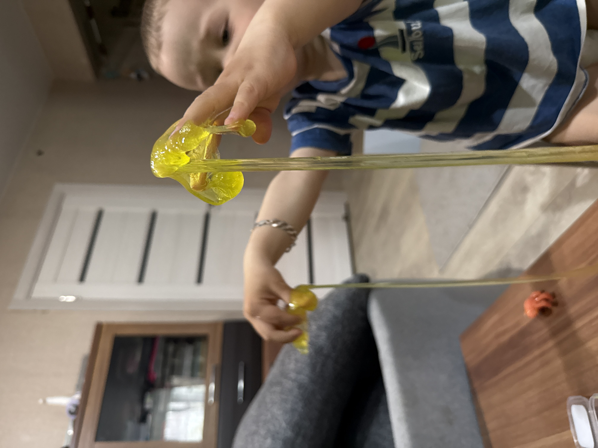 A young child wearing a blue and white striped shirt is playing with yellow slime. The slime is stretched between their hands, which are positioned above a gray sofa. A wooden table is partially visible on the right side of the image, with a small orange object on it. In the background, there is a cabinet and a white door with vertical panels. The location is likely Rusanovo, Russia.