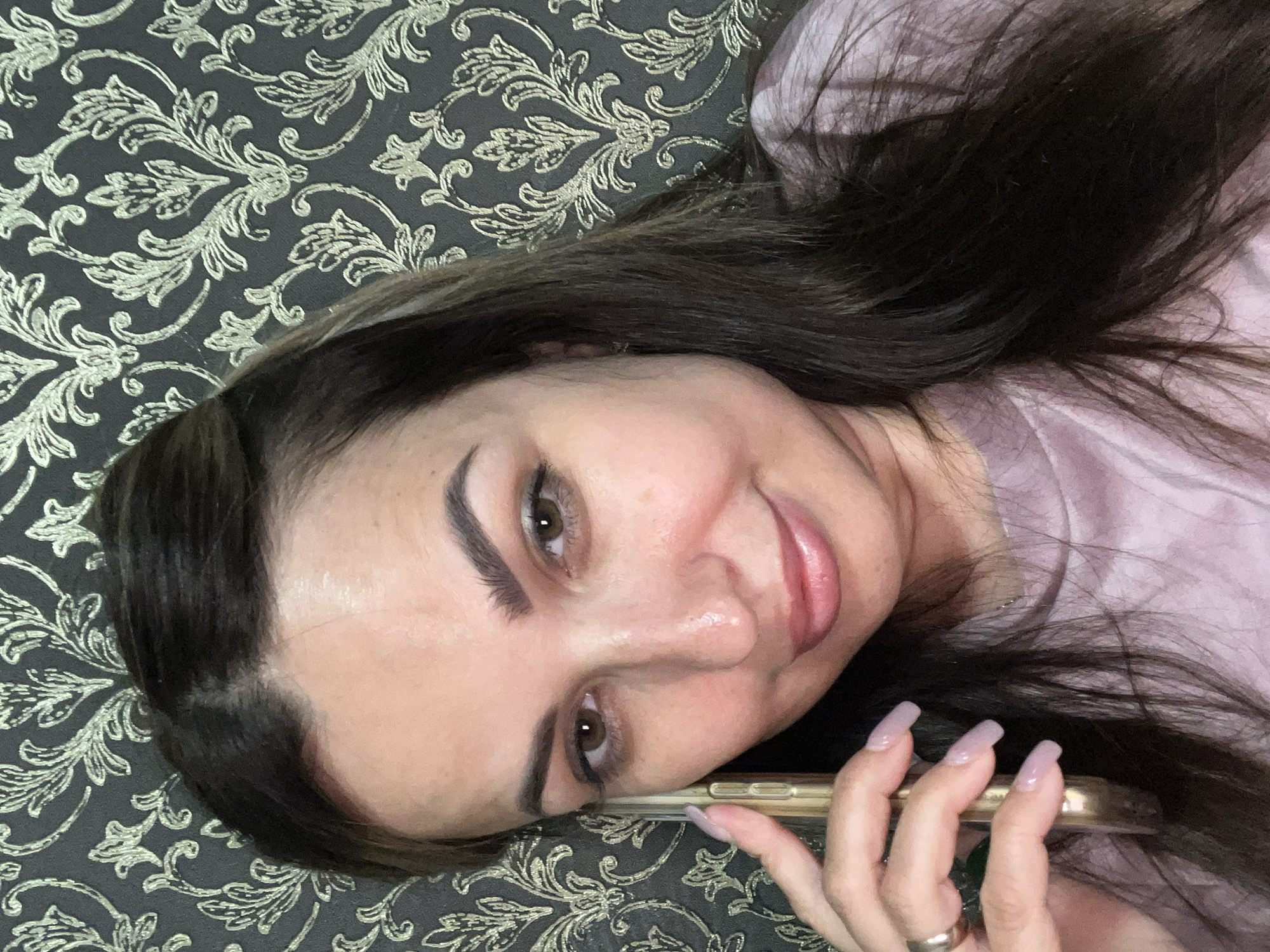 A medium close-up shot shows a woman holding a gold-colored smartphone to her right ear, with her right hand supporting the phone. The woman has dark brown hair, brown eyes, and is wearing a light pink t-shirt. She is looking at the camera with a slight smile and her head is tilted to the left. Her fingernails are long and painted a light pink color. She is wearing a gold ring on her left ring finger. The background consists of a dark gray wallpaper with a repeating ornate gold pattern. The lighting is soft and appears to be coming from the front.