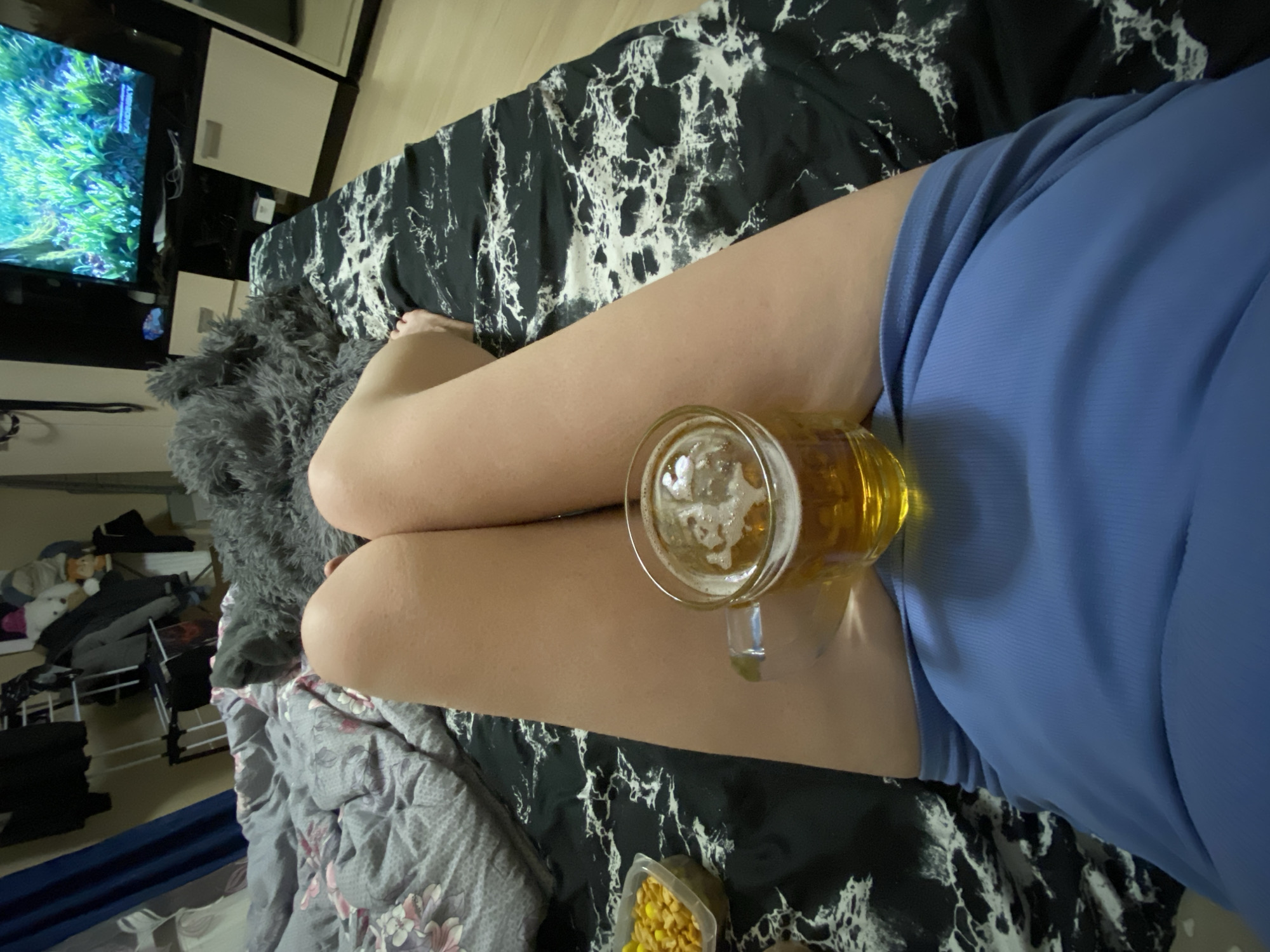 The image shows a person reclining on a bed. The person is wearing a light blue garment. On their legs rests a glass mug filled with beer. A small container of yellow snacks is also visible. The bed is covered with black and white patterned sheets and a gray blanket. A television and other furniture can be seen in the background. The location in Yekaterinburg, Russia, cannot be determined from the image.