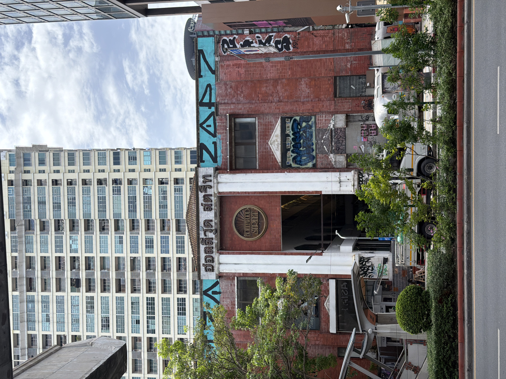 This is a street-level photograph of a multi-story building with a red brick facade, located in Bangkok, Thailand. The building features a prominent circular sign that reads "HOLLYWOOD STREET". Above this sign, there is Thai lettering and large blue graffiti that says "ZAPZ". On the right side of the building, more graffiti is visible, including black lettering. To the left of the building, a tall, light-colored apartment building dominates the background, with numerous windows and balconies. In the foreground, there is a street with a red curb, a sidewalk, and lush green bushes and trees. A white truck with a trailer is parked on the side of the building, partially obscured by vegetation. A security camera is mounted on a pole to the right of the building. The sky is partly cloudy.