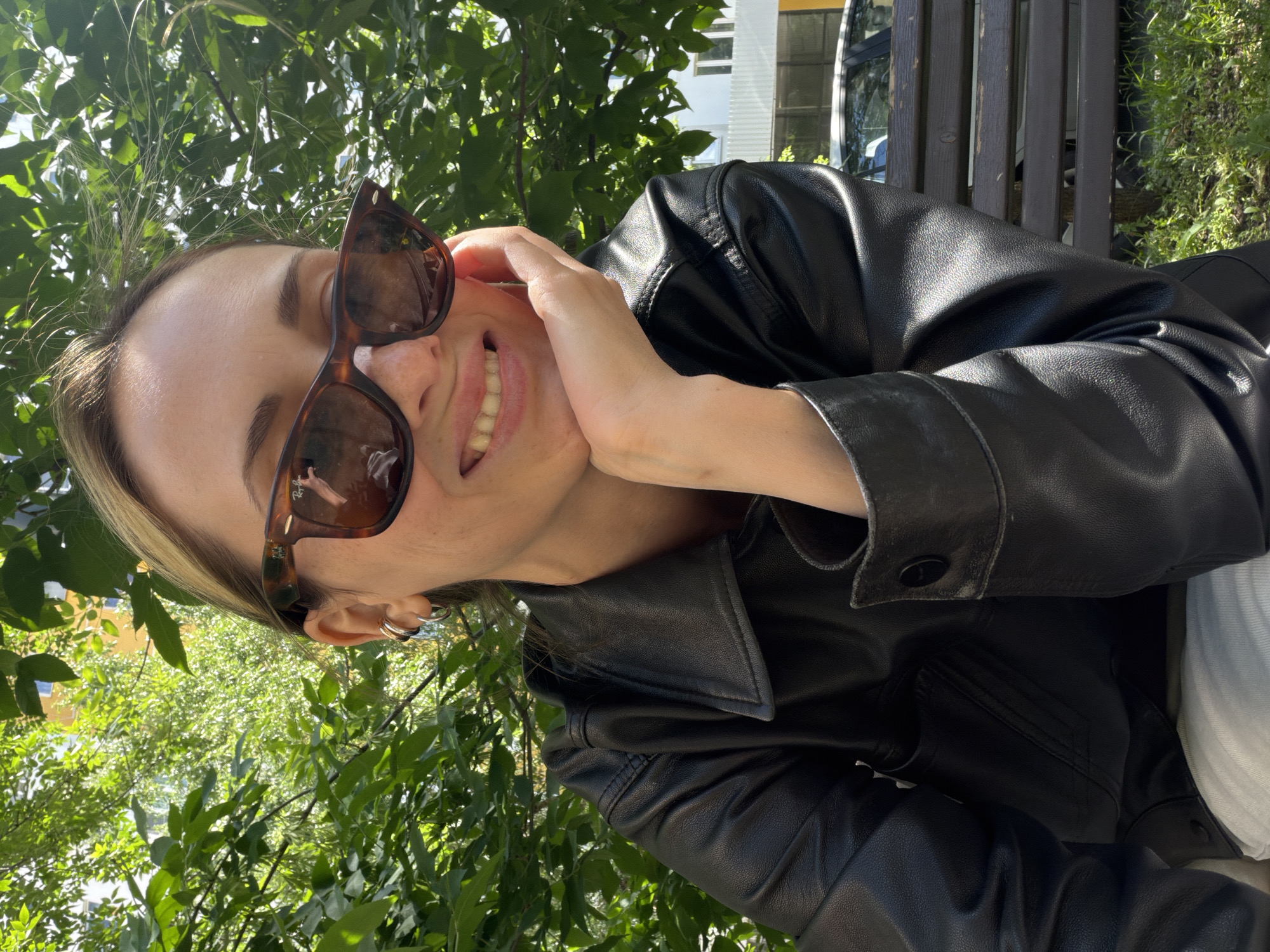 A person, oriented vertically in the frame, is captured in a close-up shot from the chest up. They are wearing a black leather jacket and dark-rimmed sunglasses. Their right hand is touching their face, near their cheek and mouth, and they have a wide smile, revealing their teeth. Green leaves and branches frame the top and left side of the image, suggesting an outdoor setting amidst nature. In the background, a wooden bench and a building with windows are partially visible, hinting at a park or a residential area. Sunlight is casting shadows, indicating it's daytime, likely sunny. The overall mood appears cheerful and relaxed. There is no visible text in the image.