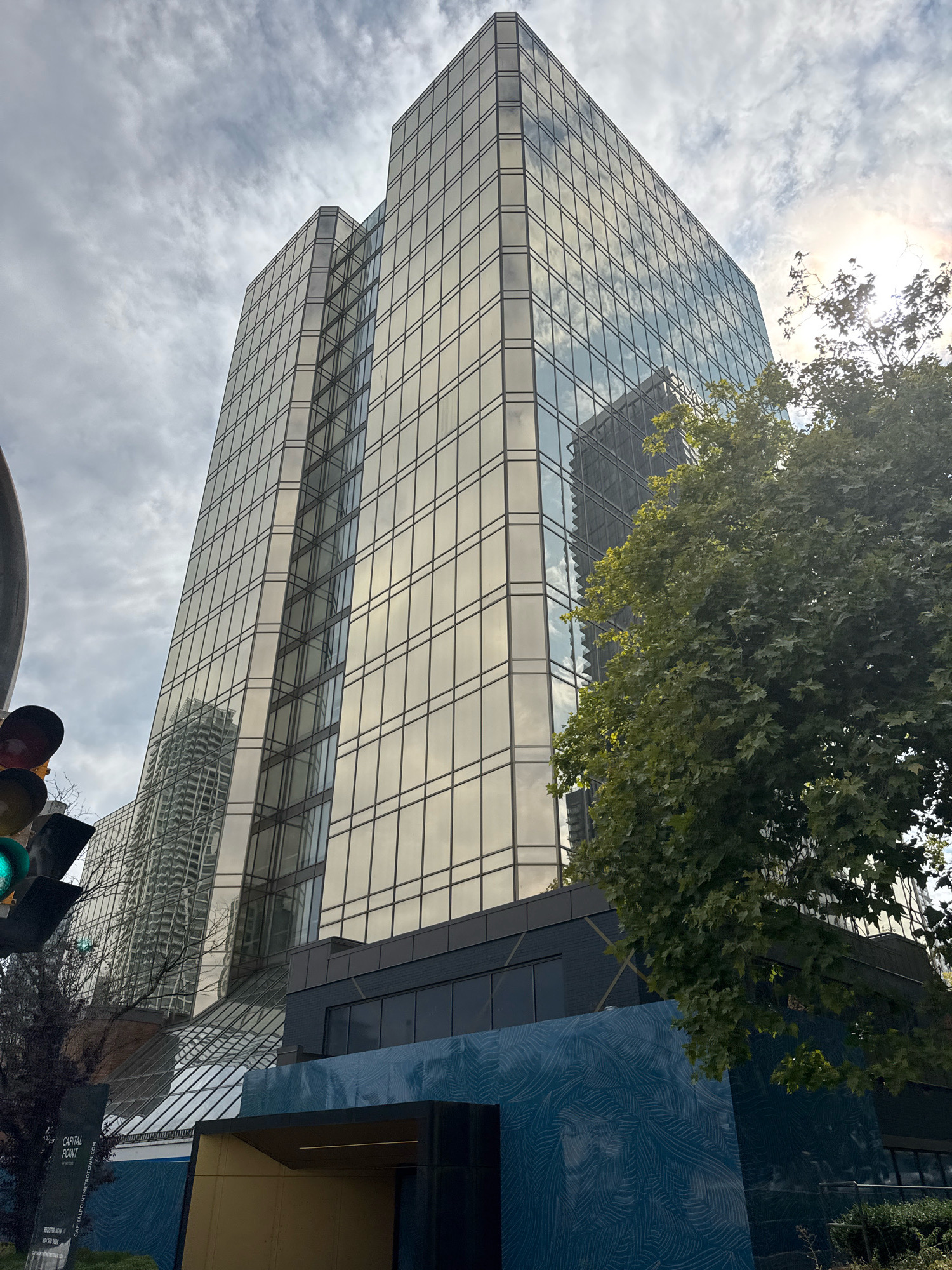 This is a low-angle, full-shot photograph of a modern, multi-story glass building in Burnaby, Canada. The building features a distinctive golden-tinted glass facade with a grid pattern of dark-colored mullions. The glass reflects the sky, clouds, and a smaller, neighboring building. To the right of the main building, a large, mature tree with lush green foliage is visible, partially obscuring the upper floors of the glass structure. In the foreground, a portion of a dark-colored lower level of the building is visible, adorned with a teal-colored graphic mural featuring a swirling, leaf-like pattern. A street sign for "CAPITAL POINT METROTOWN" is also present in the lower left corner. Traffic lights with green and yellow lights are visible on the left side of the image. The sky is overcast with scattered clouds, and the sun is peeking through the clouds on the right side, creating a lens flare effect.