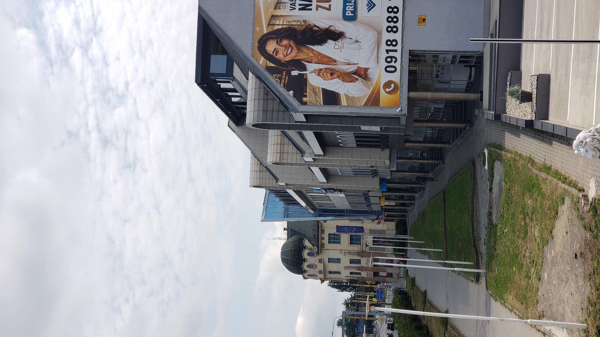 This is a medium-angle, vertical, outdoor shot showing a street scene in Prešov, Slovakia. The image is dominated by modern architecture on the right, featuring a row of multi-story buildings with a sloping, dark-colored roofline. A large billboard advertising a dental clinic is prominently displayed on the side of one building. The billboard features a smiling woman in a white coat holding a toothbrush. Text on the billboard is partially visible, including a phone number "0918 888 8".
To the left of the modern buildings, there is a more ornate, older building with a rounded, dark green dome and a pale yellow facade. This building has arched windows and is adorned with flags. The street in front of these buildings is paved with interlocking bricks and lined with grass strips and white flagpoles. A few trees are visible in the background, and traffic lights and street signs indicate it is a road intersection. One person is walking on the sidewalk in the mid-ground. The sky is overcast with scattered clouds. The foreground shows a parking lot with white lines, a small decorative planter with a potted plant, and a stone lion statue.