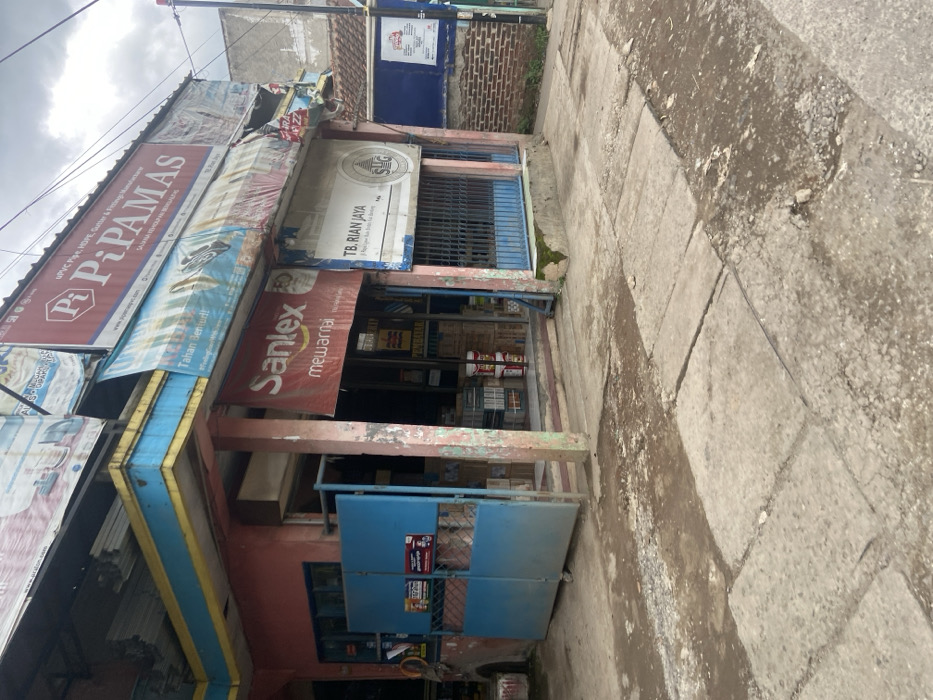 The image shows a storefront in Baleendah, Indonesia. The storefront is a single-story building with a red brick facade. There is a large, blue metal gate in front of the store, which is partially open. The storefront is adorned with various signage, including "Pi-Pamas" and "Sanlex." The storefront also displays a variety of goods, including paint cans and other building materials. The storefront appears to be located on a narrow street. The sidewalk in front of the store is cracked and uneven. The ground is covered with dirt and debris. The sky is overcast. There are no people visible in the image.