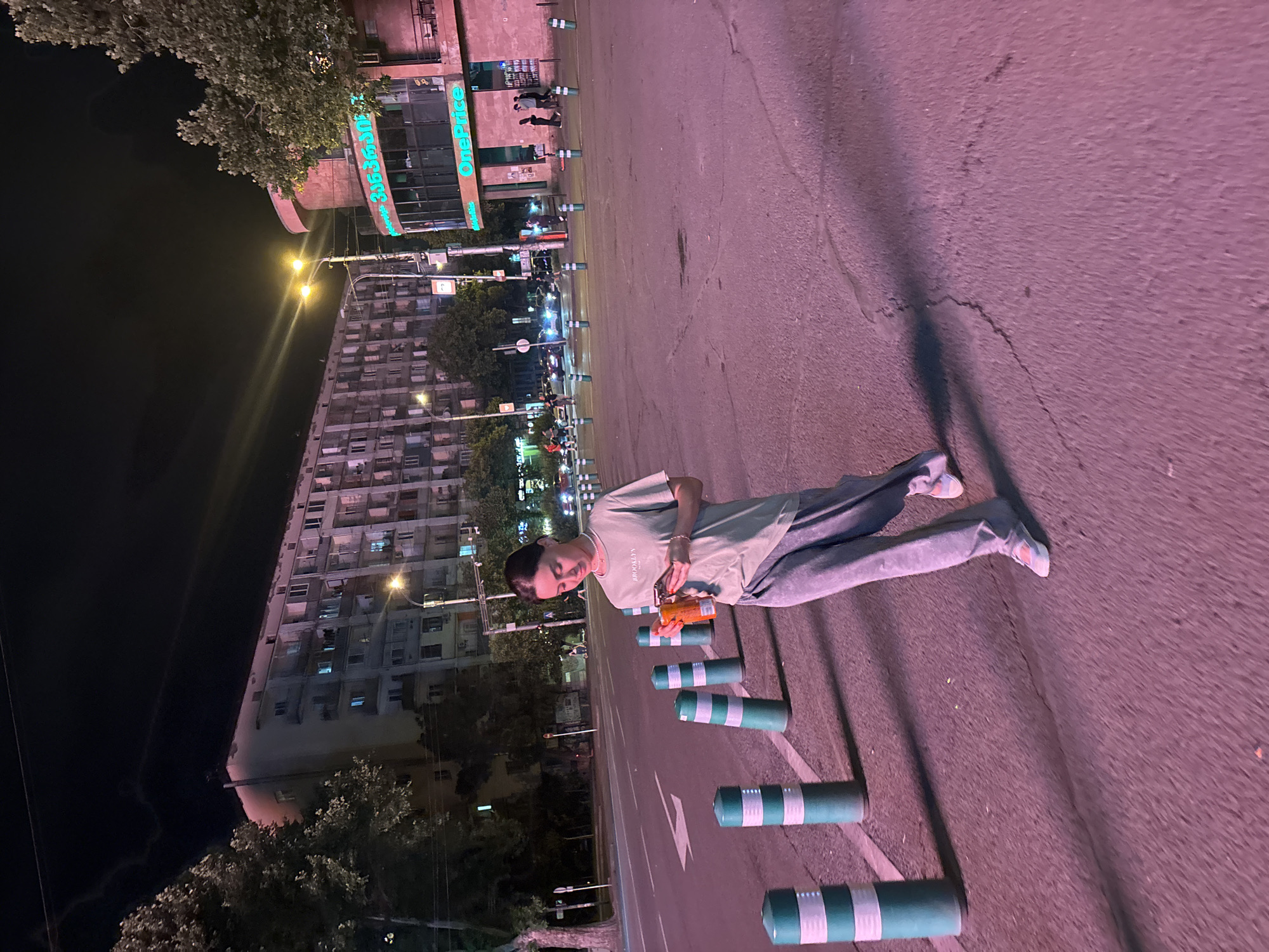 A person is lying on the asphalt of a street at night. They are wearing a light green t-shirt and grey pants. The person is holding a small orange object in their hands and looking down at it. A row of green and white bollards runs parallel to them on the street. In the background, a large, multi-story building with many windows is visible. Across the street from the building is a storefront with neon green lettering, which reads "OnePrice". Trees and streetlights are also present in the scene. The overall lighting suggests it is nighttime, and the pinkish hue of the asphalt indicates artificial lighting, possibly from streetlights or the surrounding buildings. The scene appears to be in an urban environment.