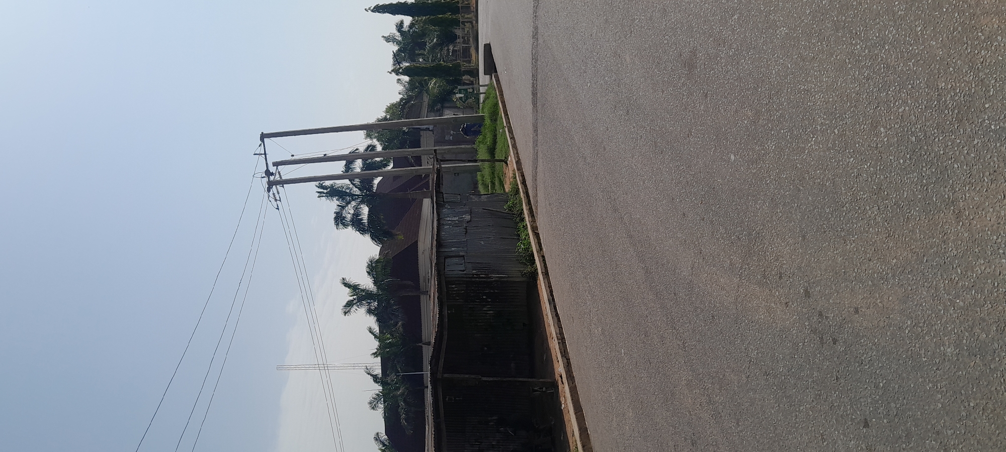 The image depicts a street scene in Jalingo, Nigeria. In the foreground, a paved road occupies the majority of the frame, curving slightly to the right. To the left of the road, a corrugated metal structure, likely a makeshift shelter or stall, is visible. Beyond the shelter, a building with a dark, sloped roof is partially obscured by palm trees. Electrical poles with overhead wires are prominent throughout the scene, indicating utility infrastructure. The sky is clear and blue. The overall environment appears to be a moderately urban or semi-rural area.