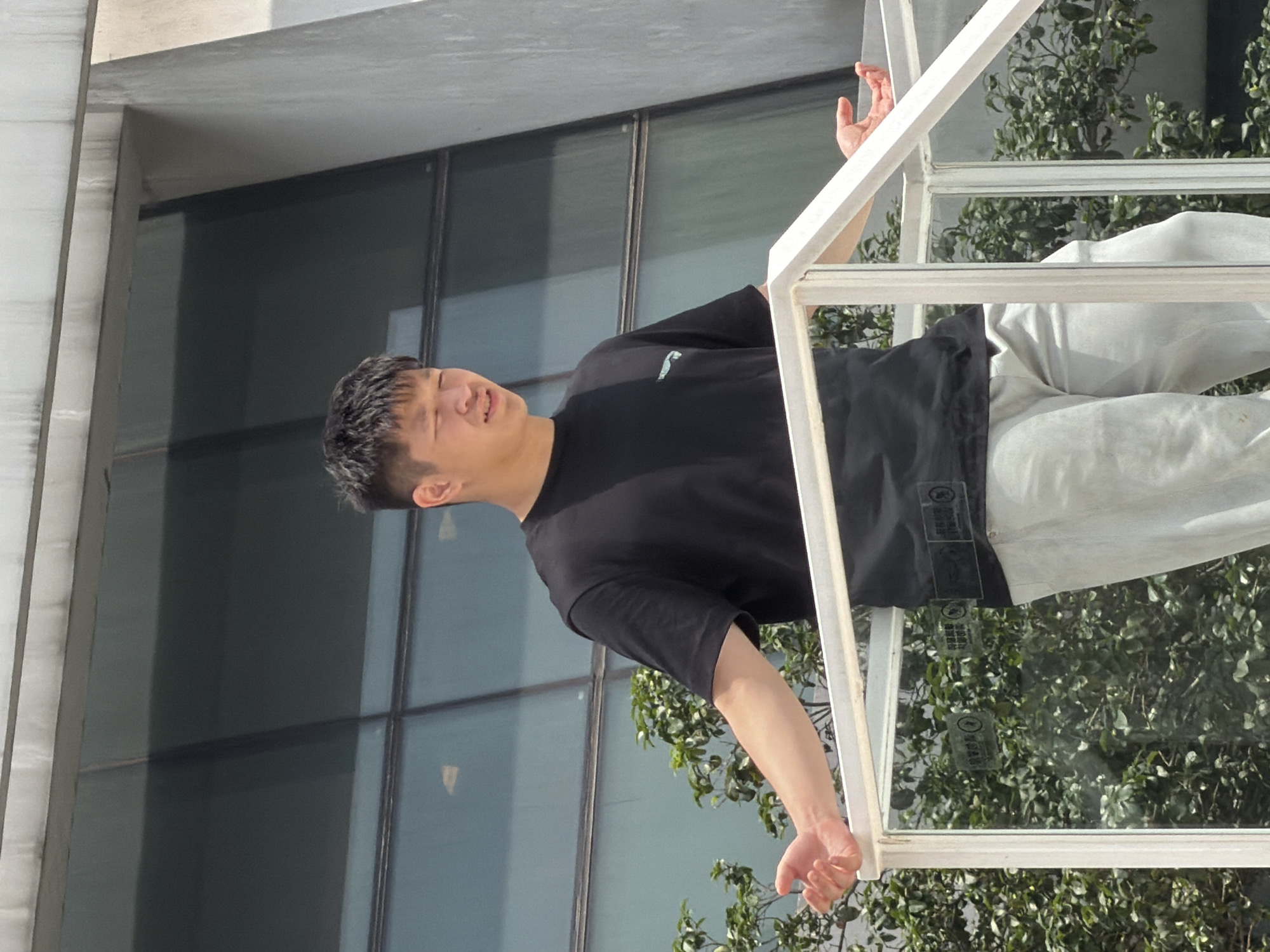 A young East Asian male stands on a raised platform in front of a modern building with large windows, possibly located in Yuzhong District, China. He is wearing a black t-shirt and light-colored pants. His arms are outstretched, and his hands are open, as if he is speaking or gesturing. He appears to be looking downwards with a slight smile. The platform has a white railing and glass panels. Lush green foliage is visible behind the glass, suggesting an outdoor or semi-outdoor setting. The lighting indicates it is daytime.