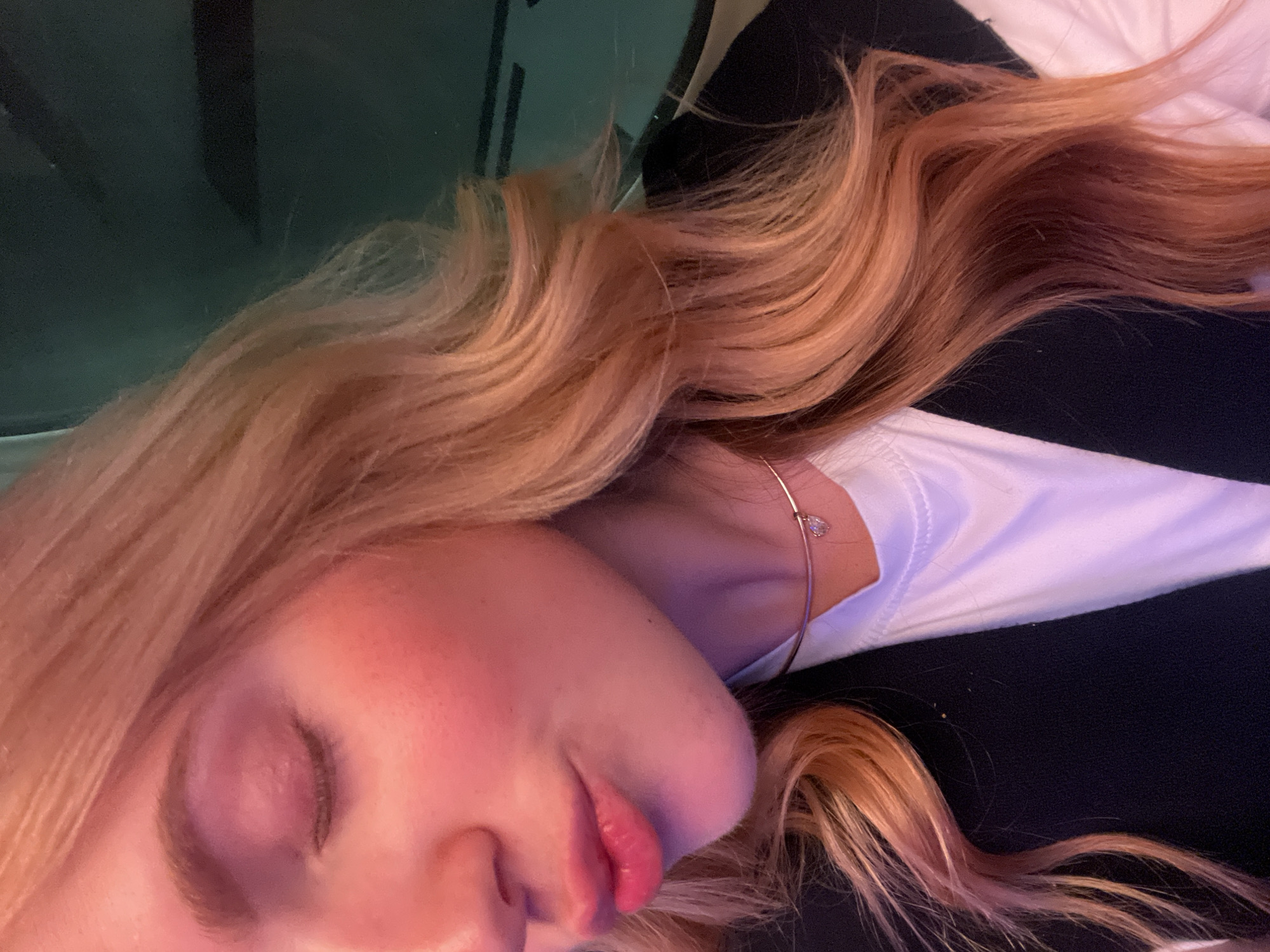 A close-up, angled view of a person lying down. The person has fair skin, closed eyes, and light pink lipstick. Their blonde hair is styled in loose waves. They are wearing a white shirt and a thin gold necklace with a teardrop pendant. The background is partially visible, with what appears to be a dark green or black surface in the upper left and a black surface to the right. No definitive details relate to the location in the city of Kempton Park, South Africa.