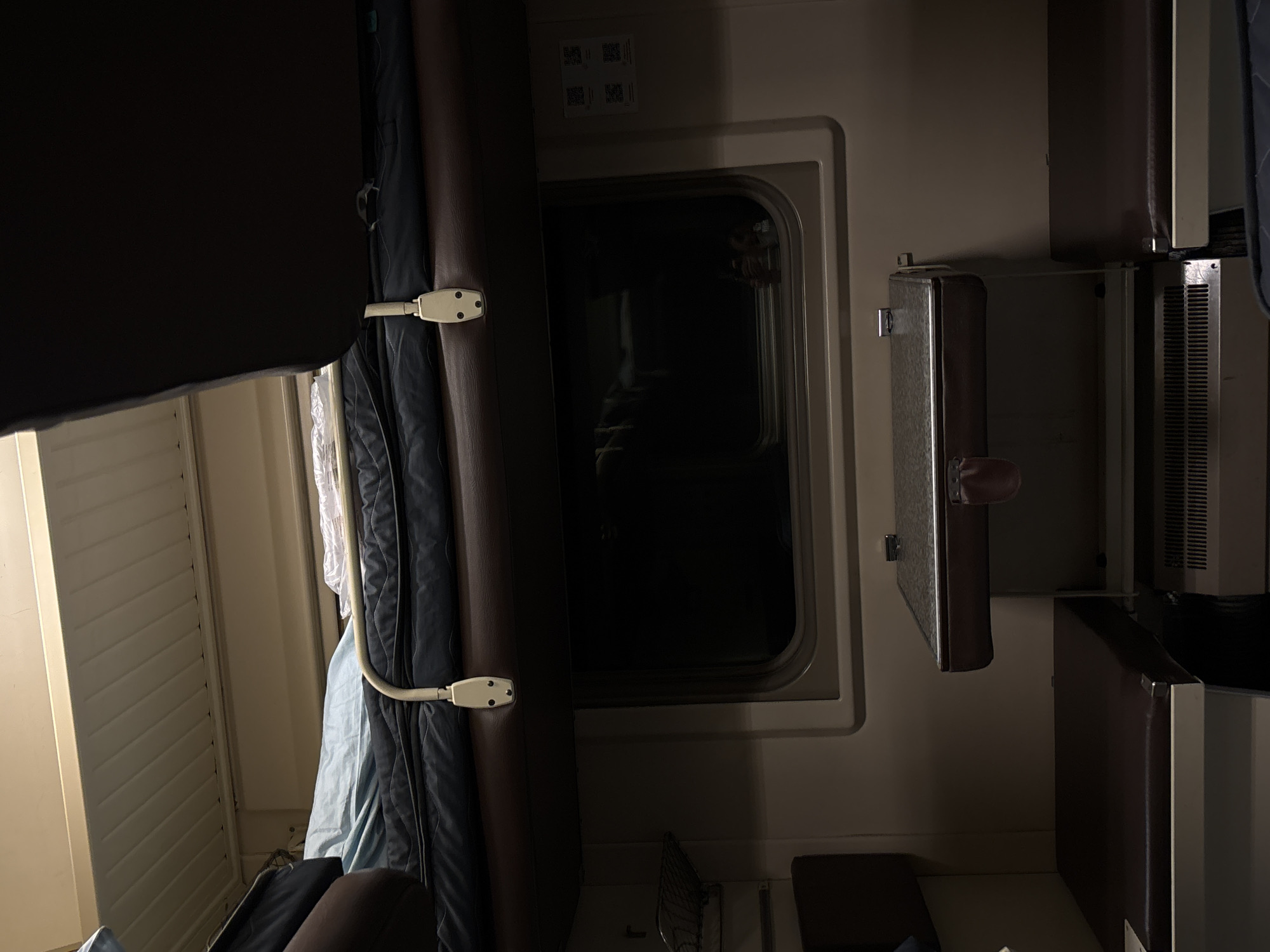 The photograph depicts the interior of what appears to be a sleeping compartment on a train. A dark window is visible, suggesting it is nighttime. A dark curtain is partially drawn, held in place by a metal rod. A luggage rack or storage compartment is mounted on the wall opposite the window. The color scheme is dominated by dark browns, blues, and grays. The location is not directly discernable from the image and cannot be identified as Шемордан, Russia, or any other specific place.