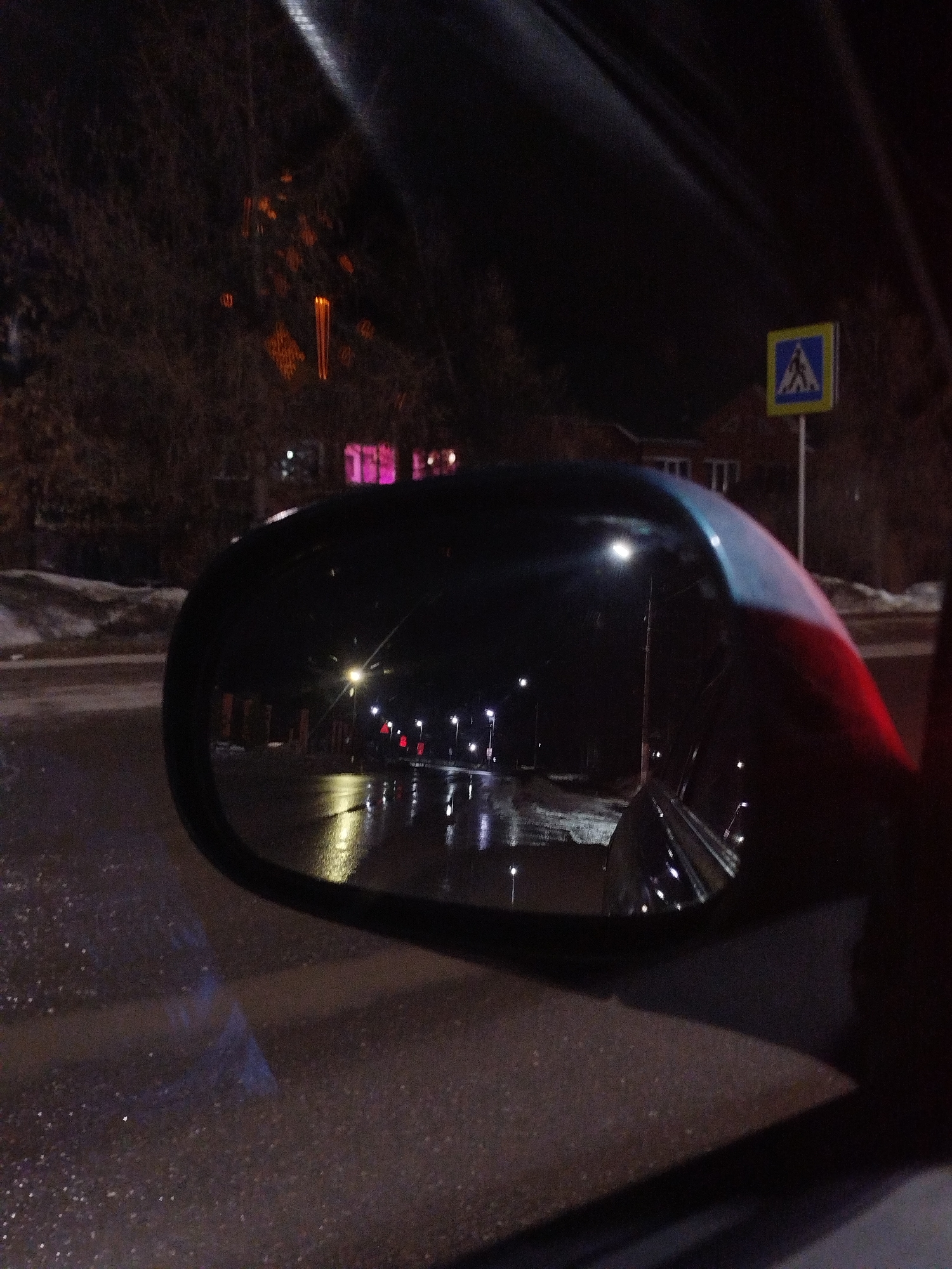 The image presents a nighttime scene observed from inside a vehicle in Chaikovsky, Russia. The immediate foreground shows a dark, wet asphalt road surface with subtle reflections. A black vehicle side mirror, likely belonging to the photographic vehicle, occupies a significant portion of the mid-right, displaying a red reflective or painted element on its outer edge. Directly visible beyond the mirror, a road extends, bordered by a snow-covered bank on the left. A standard blue square pedestrian crossing sign, featuring a white pedestrian symbol, is prominently positioned on the right side of the road on a pole. In the background, bare trees are visible, some adorned with orange and yellow decorative lights. Distant building structures are faintly discernible, with one exhibiting multiple distinctly illuminated pink windows. The side mirror's reflection reveals a straight section of road illuminated by multiple streetlights, casting elongated, bright reflections on the wet asphalt surface. No individuals are visible in the scene.