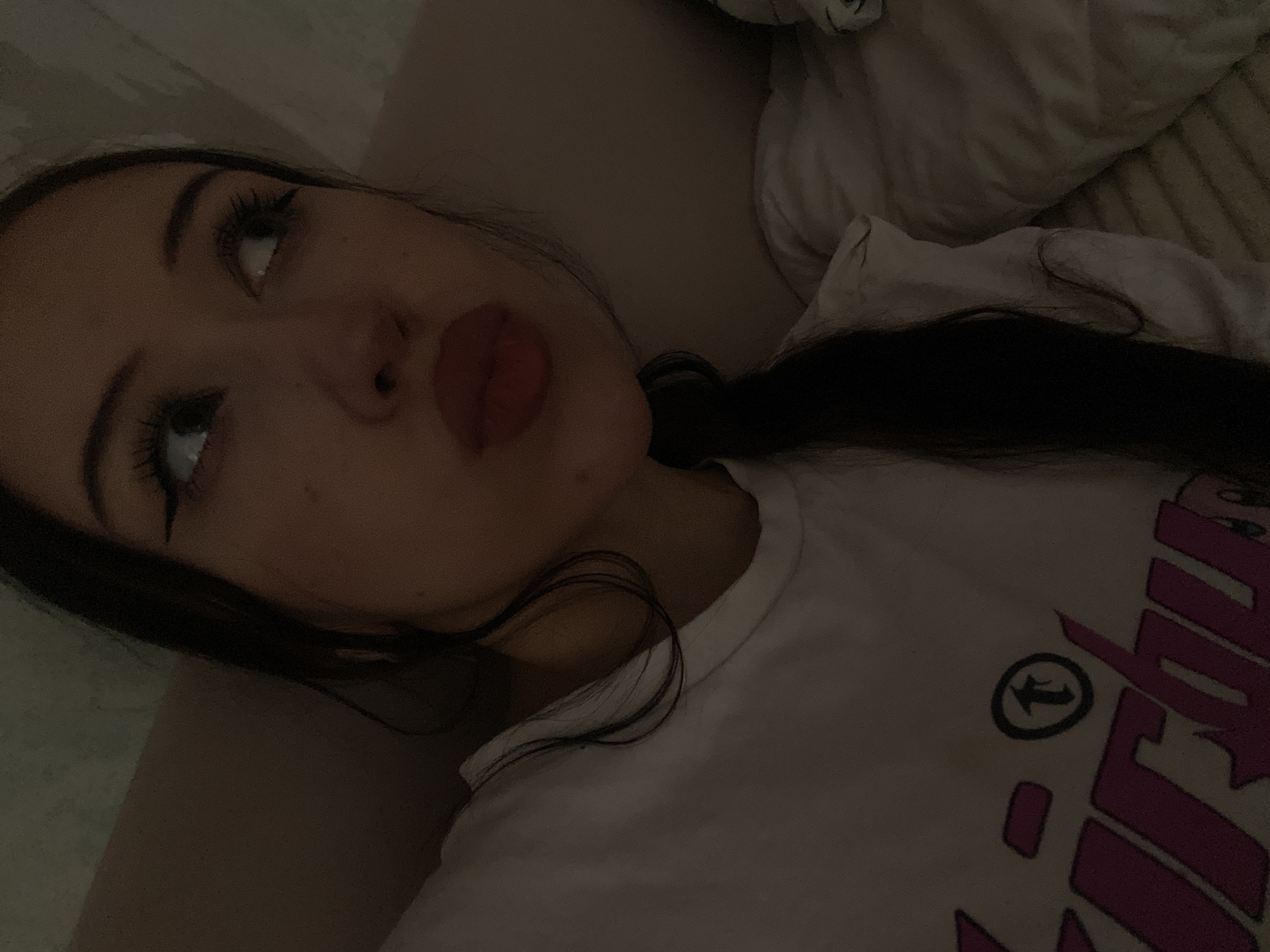 The media file is a close-up, overhead shot of a young woman lying down in what appears to be a bed. The lighting is dim, suggesting either nighttime or a room with little natural light. The woman is looking upwards and to her left, with a slight pout on her lips. Her eyes are made up with dark eyeliner and mascara. Her dark hair is spread out around her. She is wearing a white t-shirt with a graphic design on it. The design includes large, stylized pink lettering and a circular logo with an arrow inside. The background consists of pillows and bedding, further indicating a bedroom setting. The overall mood conveyed by the image is relaxed and perhaps a bit introspective due to the subject's gaze and the dim lighting. There are no explicit cues to the time of day or weather. The location context provided, Votkinsk, Russia, is not visually evident in the image itself.