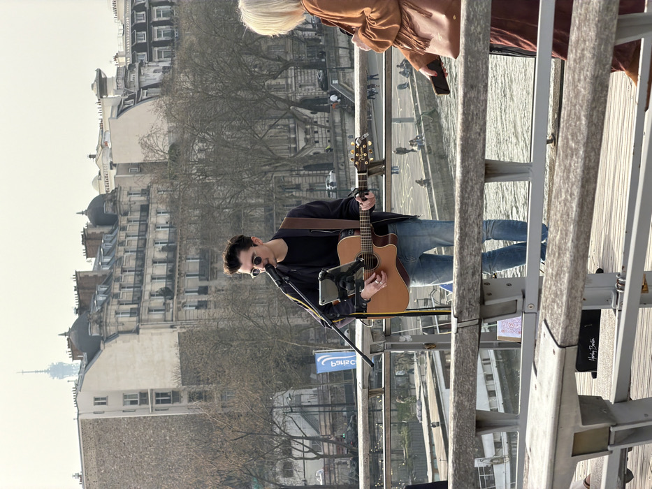 The scene captures a daytime outdoor performance on a bridge or elevated walkway in Paris, France, with the Eiffel Tower discernible in the distant background.
A young adult male musician, wearing sunglasses, a dark jacket, dark t-shirt, and light blue jeans, is centrally positioned, standing and actively playing a natural wood acoustic guitar. He is singing into a microphone attached to a stand, which also holds a tablet device. An amplifier, marked "Harley Benton," rests on the ground near his feet.
To the right, partially obscured by a wooden and metal railing that stretches across the foreground, is a person with blonde hair, dressed in a brown fringed jacket and carrying a brown shoulder bag. This individual holds a smartphone and appears to be observing the musician or the river.
Below the walkway, the Seine River flows, its surface reflecting light. Stone quays are visible along the riverbanks, with a few individuals present and a boat moored. In the background, characteristic Parisian buildings with balconies and distinct rooftops line the river, interspersed with numerous leafless trees. The Eiffel Tower stands prominently in the hazy distance, slightly left of center, beneath a pale sky.