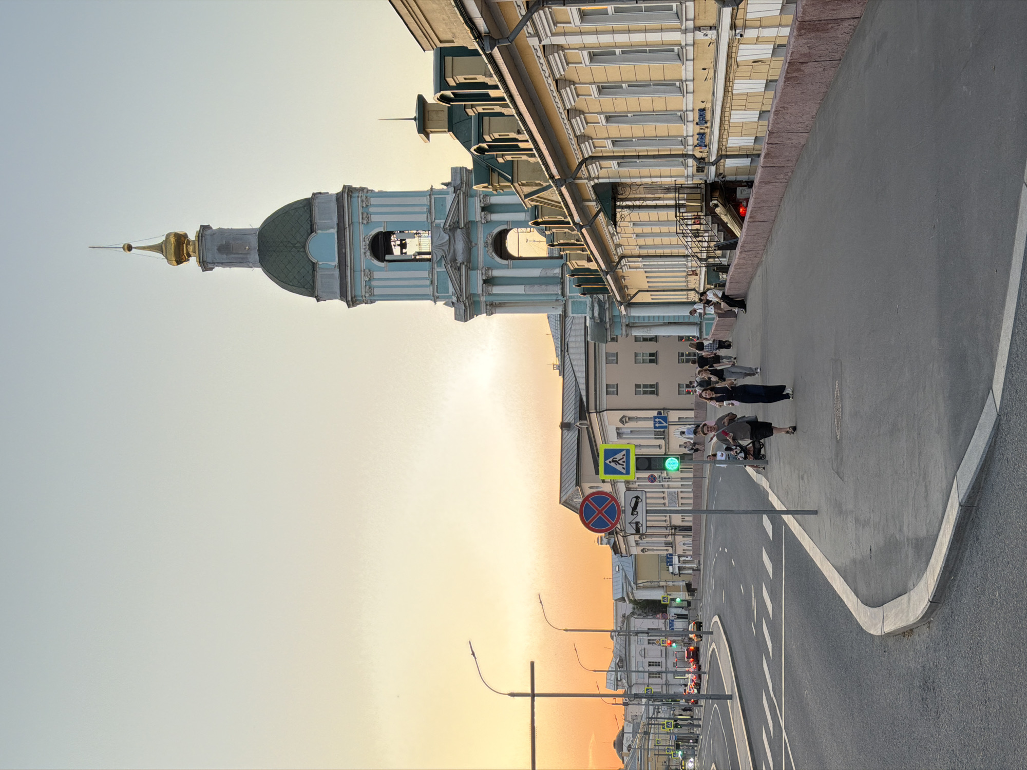 The image depicts a cityscape in Moscow, Russia. The prominent architectural feature is the Naval Cathedral of Saint Nicholas, characterized by its blue facade and golden dome. Adjacent to the cathedral is a multi-story building with a pale yellow exterior.
A group of approximately 5-7 individuals are standing on a raised pedestrian walkway. The walkway is bordered by a road with zebra-striped pedestrian crossings and street lights. There are multiple traffic signs visible, including a pedestrian crossing sign and a no-parking sign.
The overall scene suggests a street view during the late afternoon or early evening, as evidenced by the warm, golden hue in the sky.