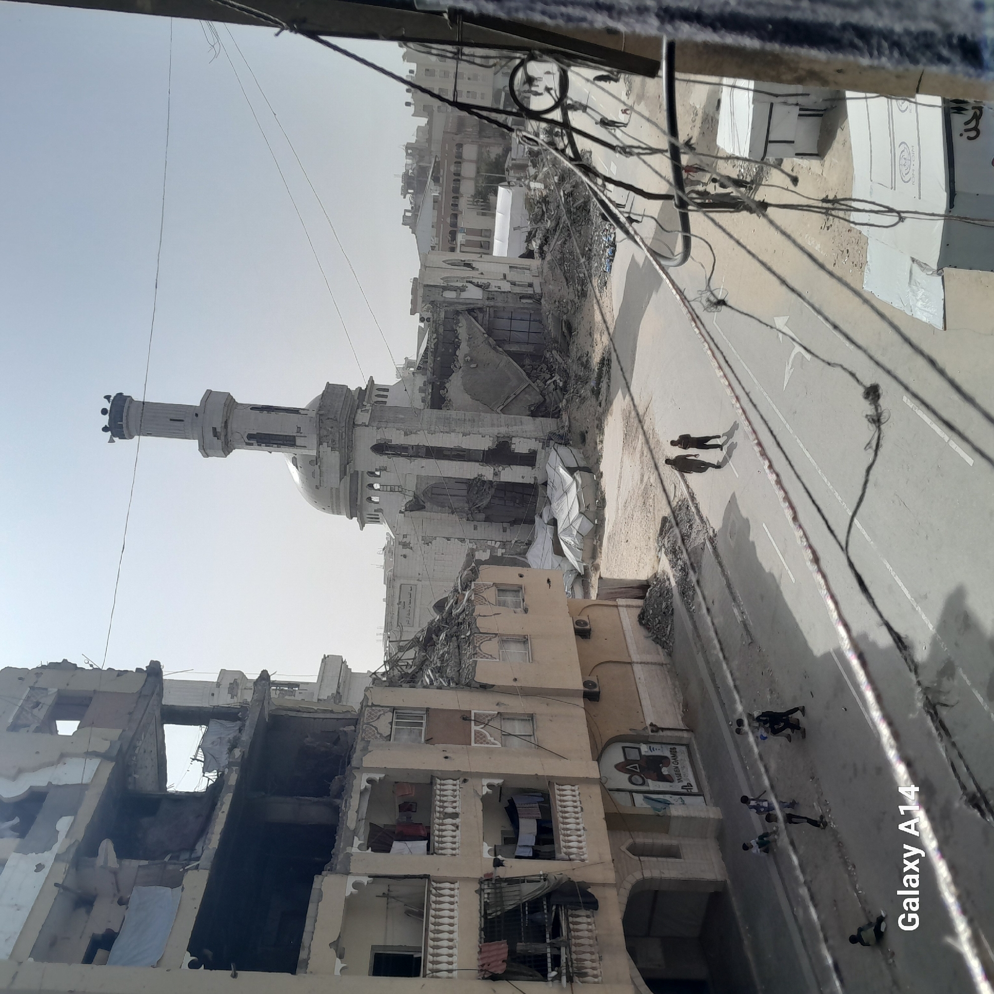 This image depicts a street in an urban environment, possibly the aftermath of conflict. There are damaged buildings with visible destruction, including one appearing to be a mosque with a minaret. Two individuals are visible walking along the street, while a larger group of people are present further down the street. Overhead, there are numerous wires and cables. The overall scene conveys an atmosphere of devastation. The image includes the text "Galaxy A14". The location appears to be in the city of Unknown, Unknown.