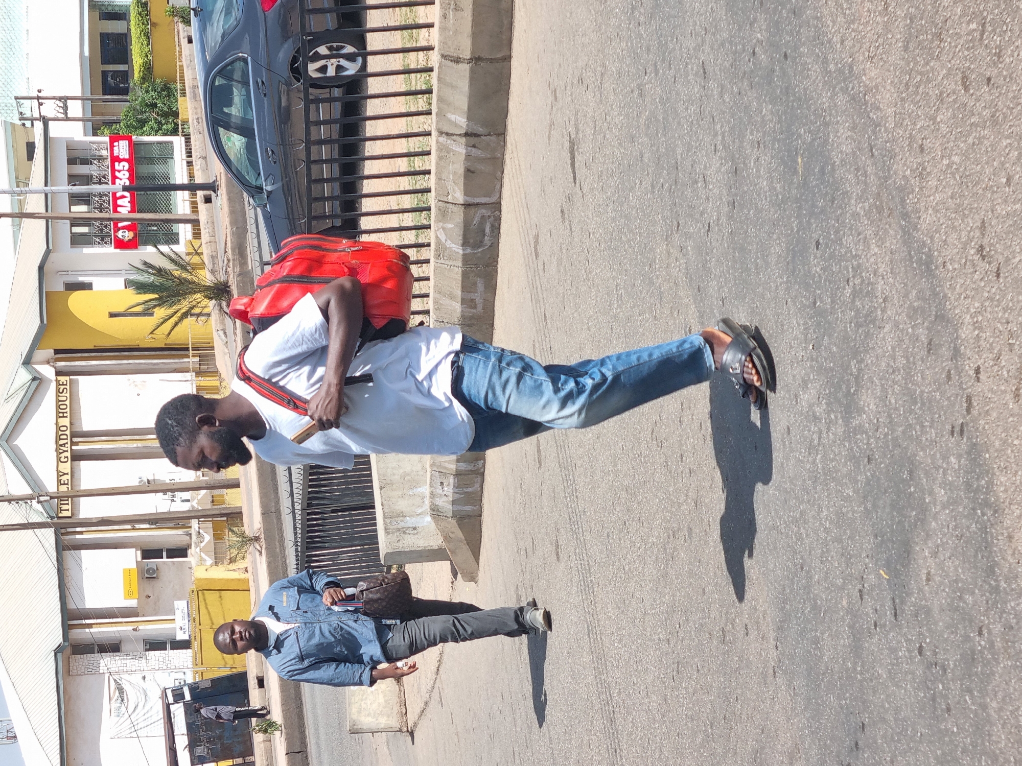 A street scene in Jos, Nigeria shows two men. The first man is Black with a beard. He wears a white T-shirt, blue jeans, and black sandals. He carries a red backpack. The second man is also Black. He wears a blue denim shirt, dark pants, and grey shoes. He carries a brown handbag. They are standing on a street. A black car is parked near a fence. A building with a sign that reads "Tilly Gyado House" is in the background. A red sign with "365" is visible on a building across the street.