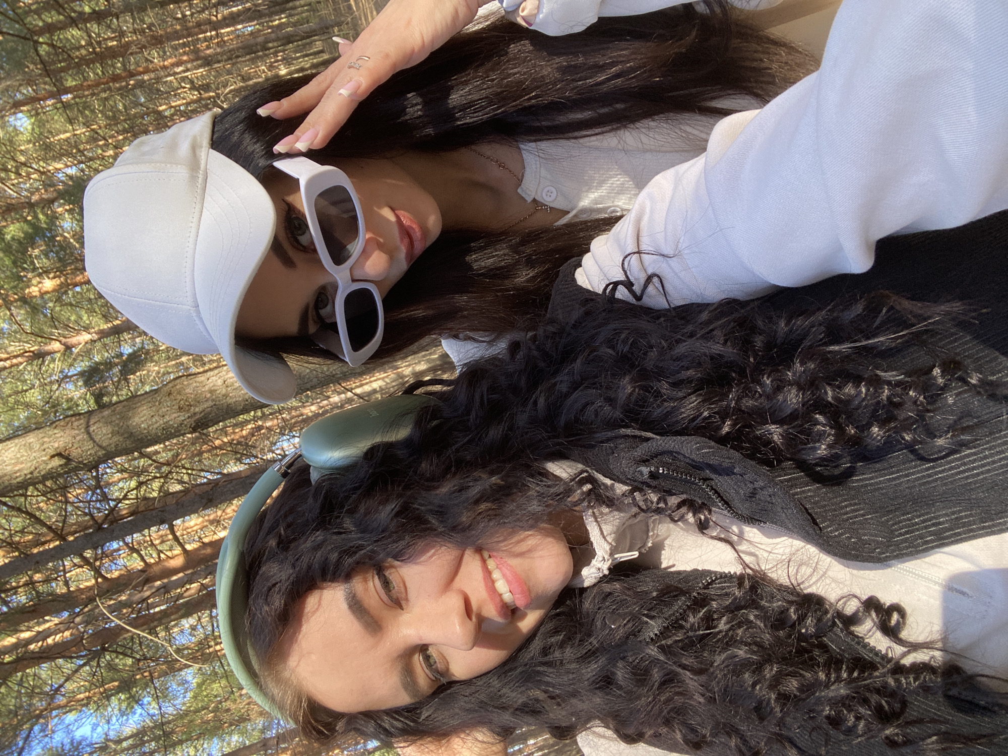 Two women are captured in a selfie against a backdrop of tall trees, likely in a forest or wooded area. The background suggests a sunny day with dappled light filtering through the trees.
The first woman, positioned at the top of the frame, is wearing a white baseball cap and oversized white sunglasses. She has long, dark hair. Her nails are manicured with a French tip design. She is wearing a white sweater.
The second woman, at the bottom of the frame, has dark, curly hair and green over-ear headphones.
The location is not specified in the image and is not verifiable using visual elements alone. It is not possible to confirm the location is in Chaikovsky, Russia, based on the visual information provided.