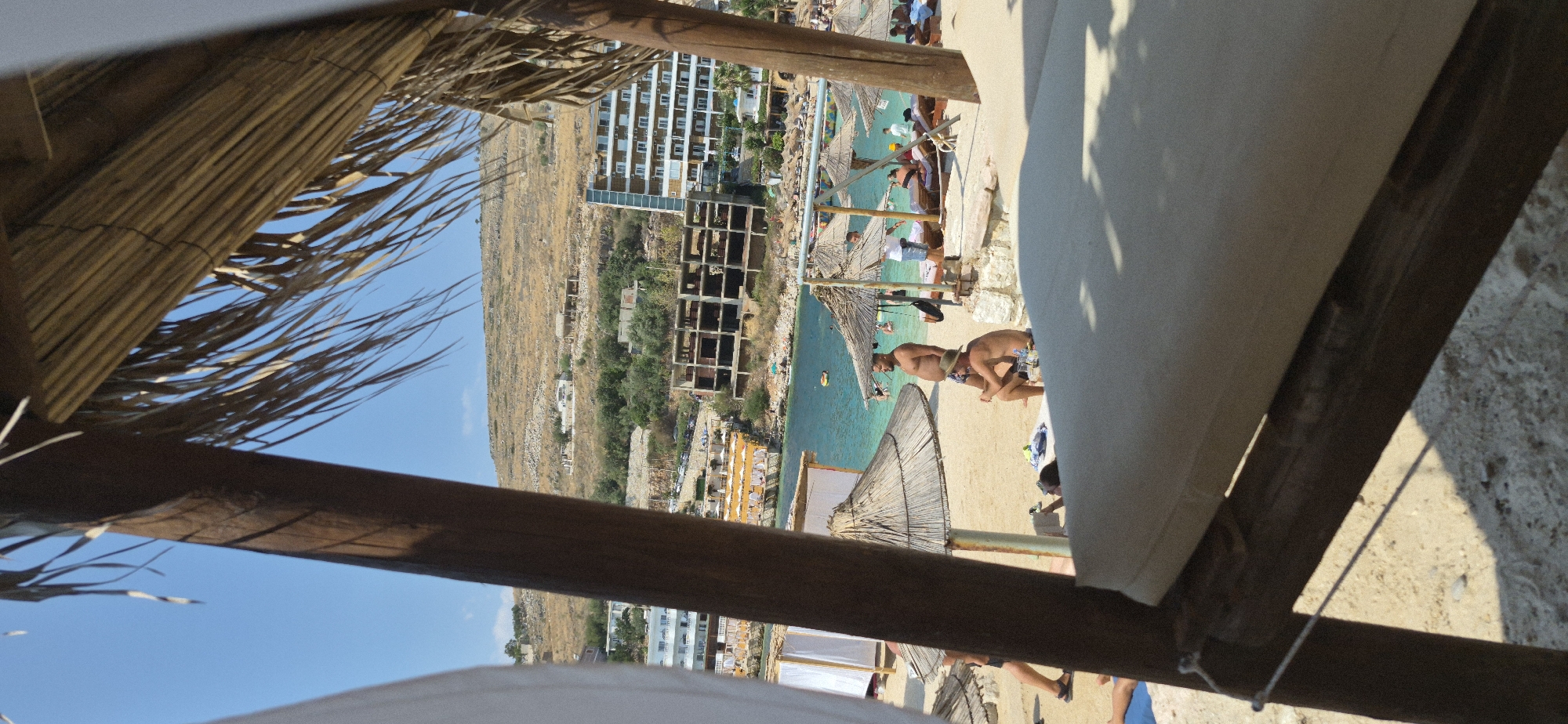The image captures a beach scene in Saranda, Albania, under a clear blue sky. In the foreground, a large wooden post and a thatched roof create a partial frame, suggesting a beach umbrella or shelter. The sand is visible, with scattered rocks and shadows cast by the overhead structure.
Further back, on the sandy beach, several people are present. Two men are prominently seated under a thatched umbrella. One man, wearing a hat, is sitting while the other, who is larger and shirtless, stands. Other individuals can be seen lying on lounge chairs or walking on the beach.
Beyond the beach, the turquoise waters of the Ionian Sea are visible, with a few small boats or inflatable toys in the water. Along the coastline, a resort area is apparent, featuring buildings with multiple stories, some appearing to be hotels or apartment complexes. There are also several unfinished or under-construction buildings, indicating ongoing development. Further inland, a hillside rises, dotted with smaller buildings and vegetation. The overall impression is of a lively beach resort on a sunny day.
