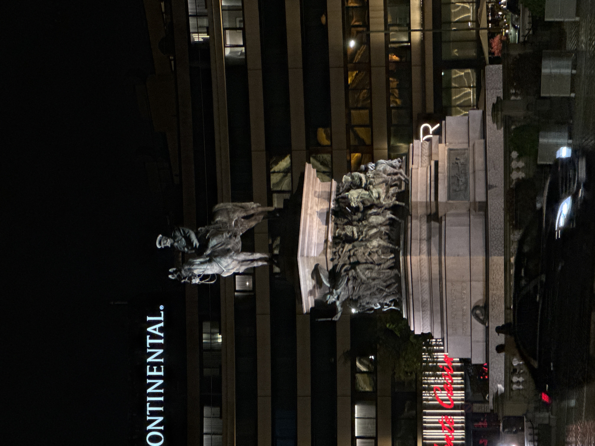 At night, the image shows a vertical view of the Equestrian Monument to Tsar Liberator Alexander II in Sofia, Bulgaria. Behind the monument, the word "CONTINENTAL." is lit vertically on a building, and a red sign is partially visible near the base of the monument. A vehicle is on the right, with its headlights reflecting. The surrounding environment appears urban.