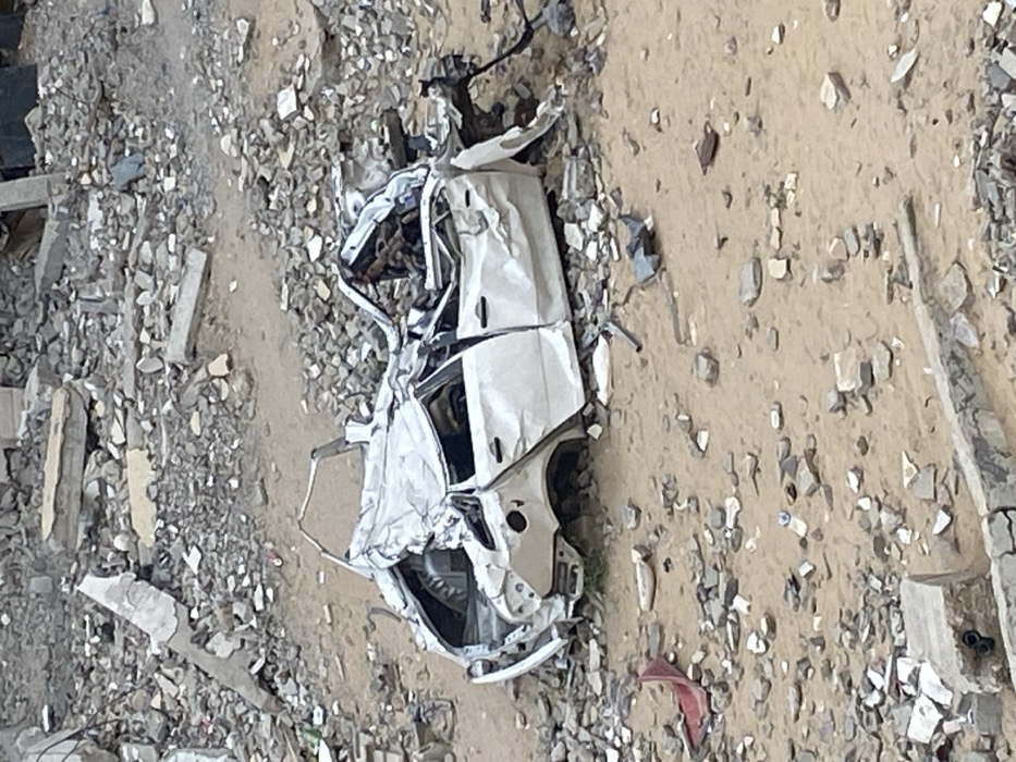 The image shows a severely damaged white car amidst debris. The car's frame is twisted, and parts of the body are crumpled. The surrounding area is covered in rubble, with a mix of gravel, concrete fragments, and dirt. The destruction suggests a catastrophic event, possibly an explosion or a collision. The car's mangled state indicates a high impact force.